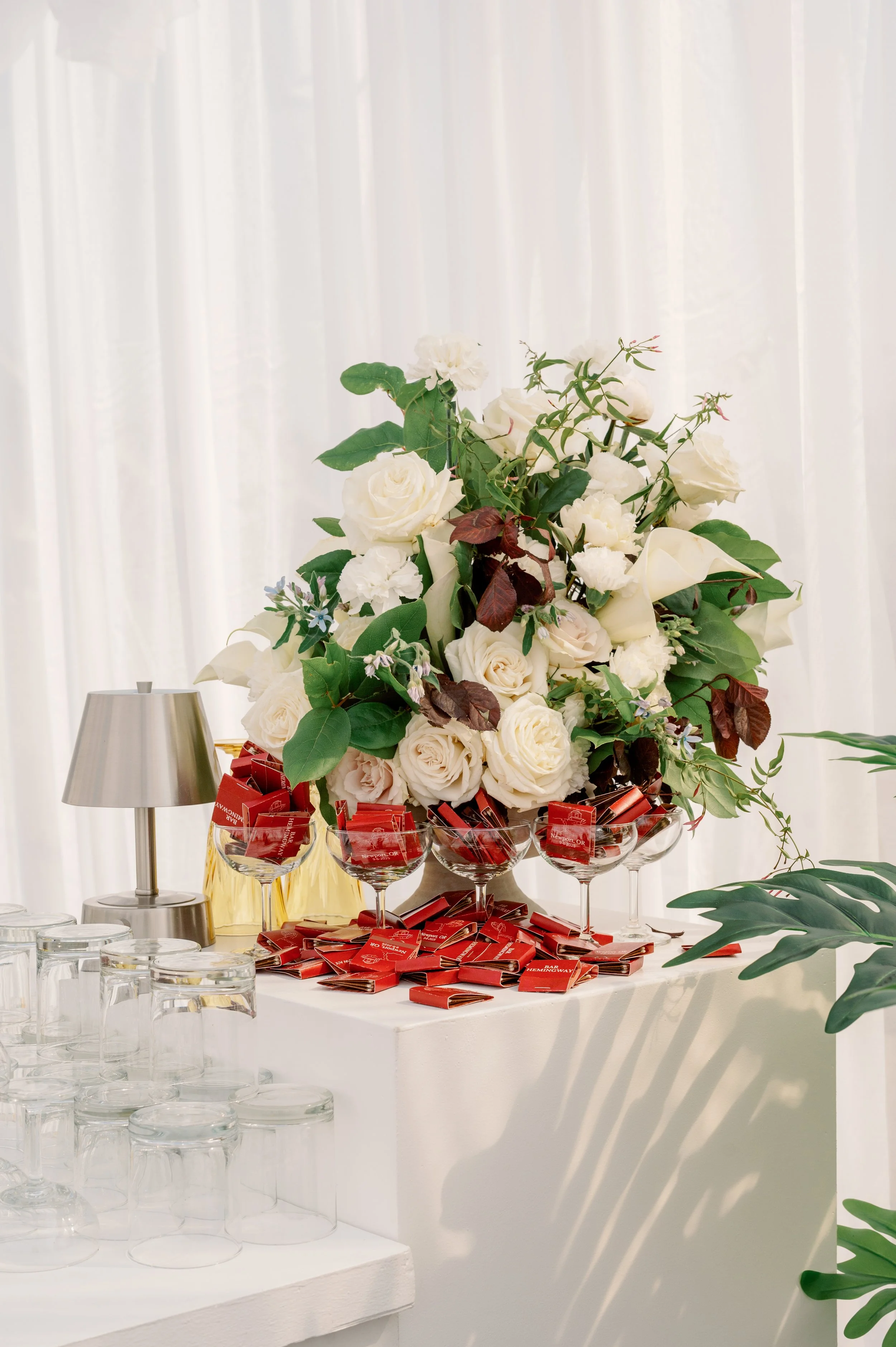A floral arrangement of white roses and other greenery on a table, decorated with red wrapped chocolates, next to glassware and a silver lamp, with a white curtain background.