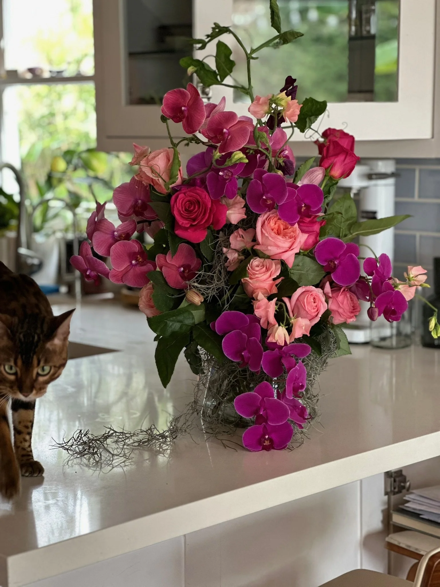 A colorful flower arrangement with pink roses, purple and pink orchids, and other small flowers in a glass vase on a kitchen counter, with a window in the background showing greenery outside.