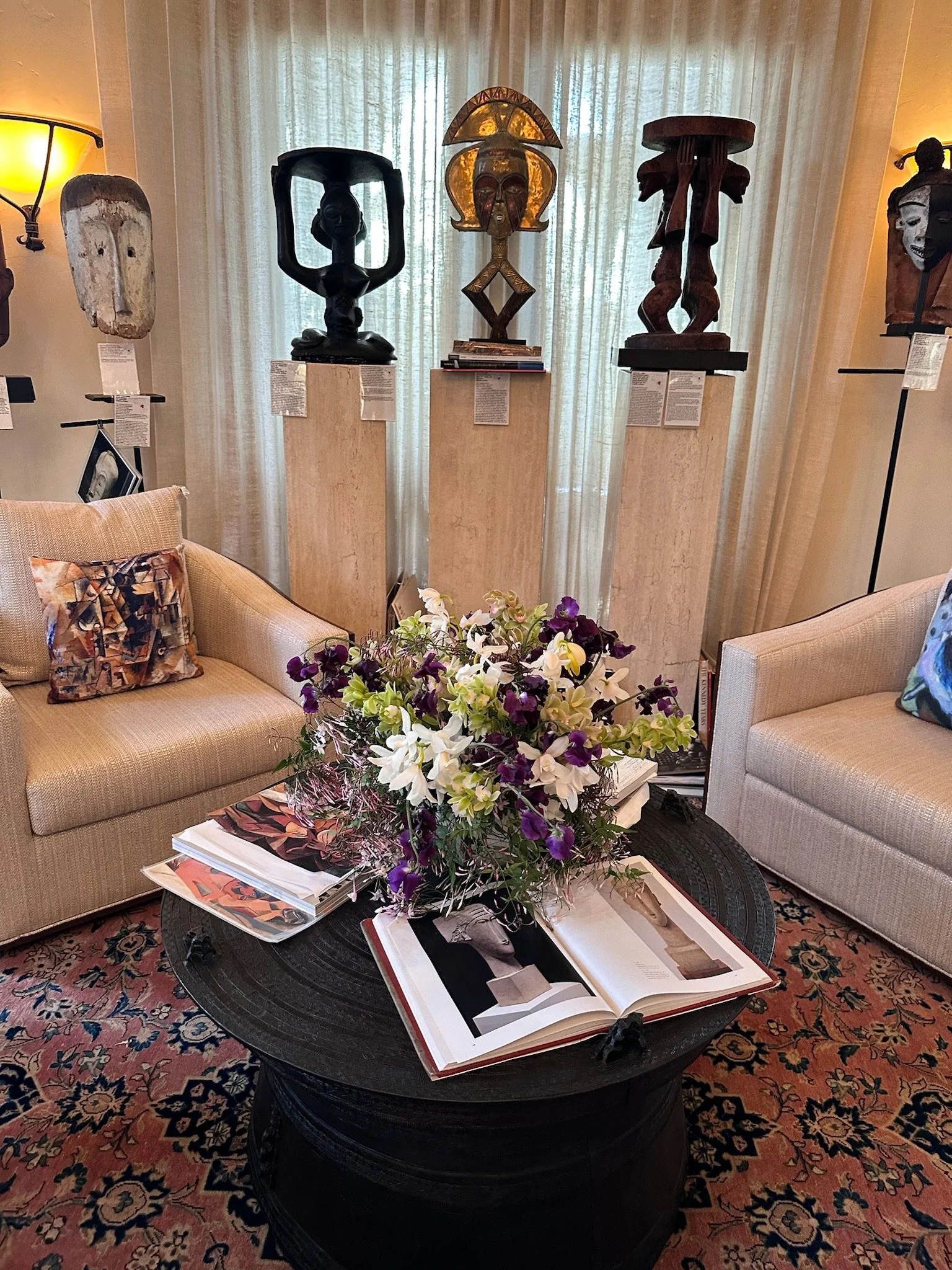 Living room with a dark oval coffee table with a large bouquet of white and purple flowers, open magazines, and books. There are two beige sofas with colorful throw pillows, a patterned carpet, and a textured area rug. In the background, four African art masks are displayed on pedestals with informational placards. The curtains are light-colored, and warm ambient lighting is from lamps on the walls.