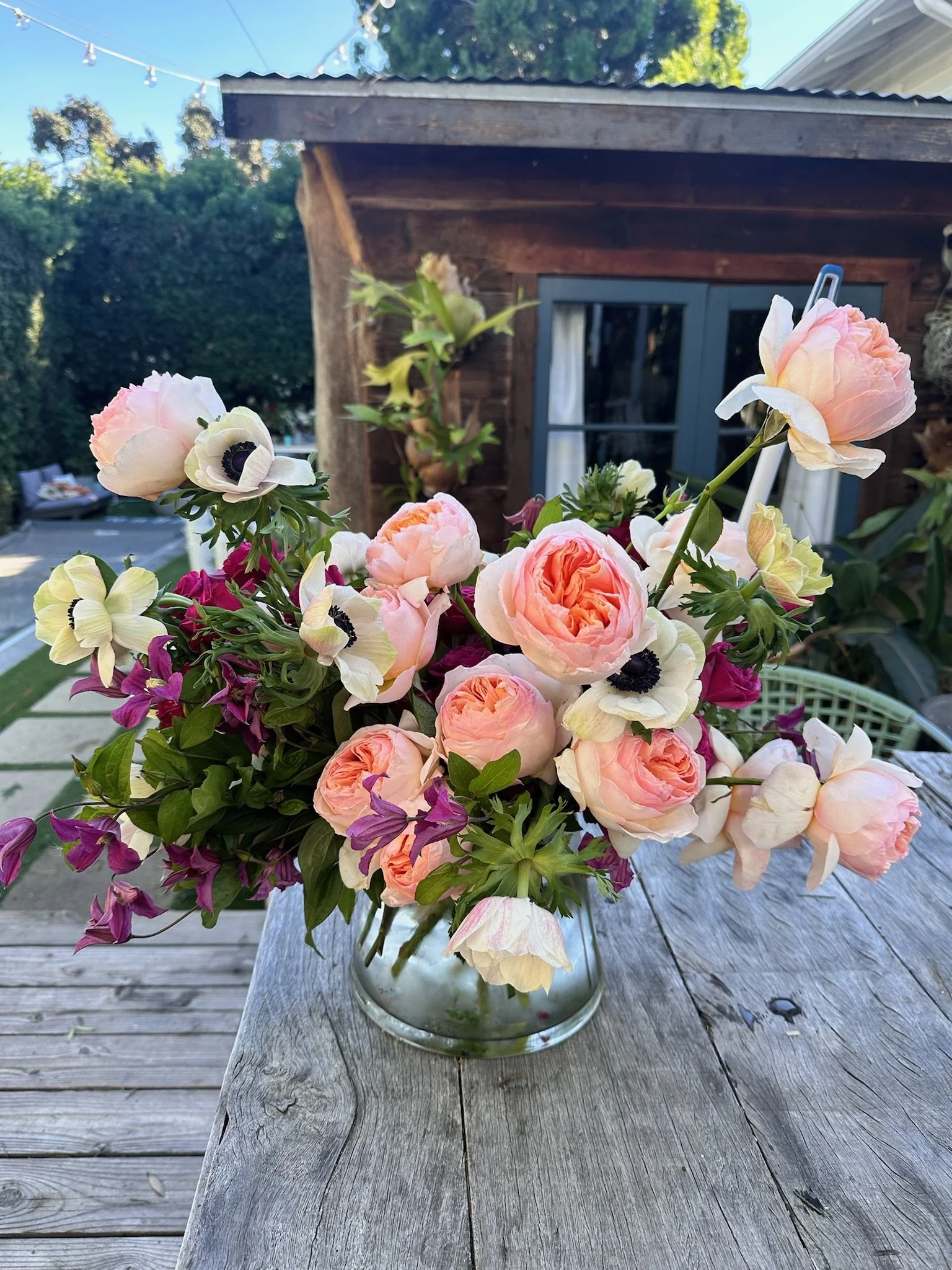 A glass vase filled with a colorful bouquet of assorted flowers, including pink roses, white anemones, and purple and yellow flowers, placed on a weathered wooden table outdoors.