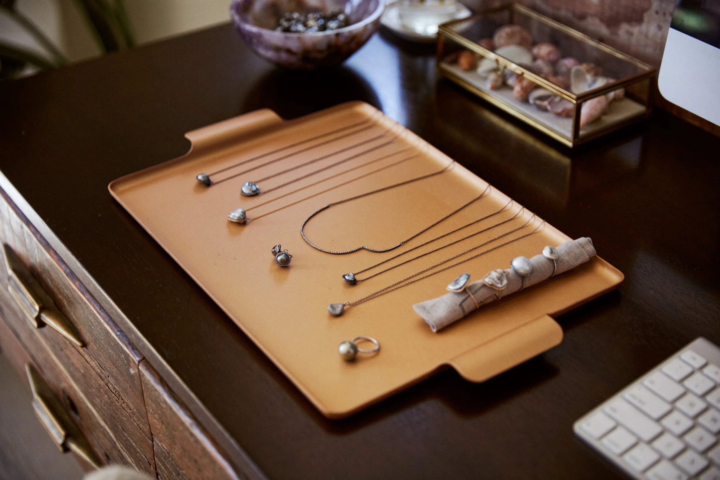 Jewelry displayed on a tan tray, including necklaces, rings, and earrings, with a bowl of jewelry and a glass jewelry box in the background on a dark wooden surface.