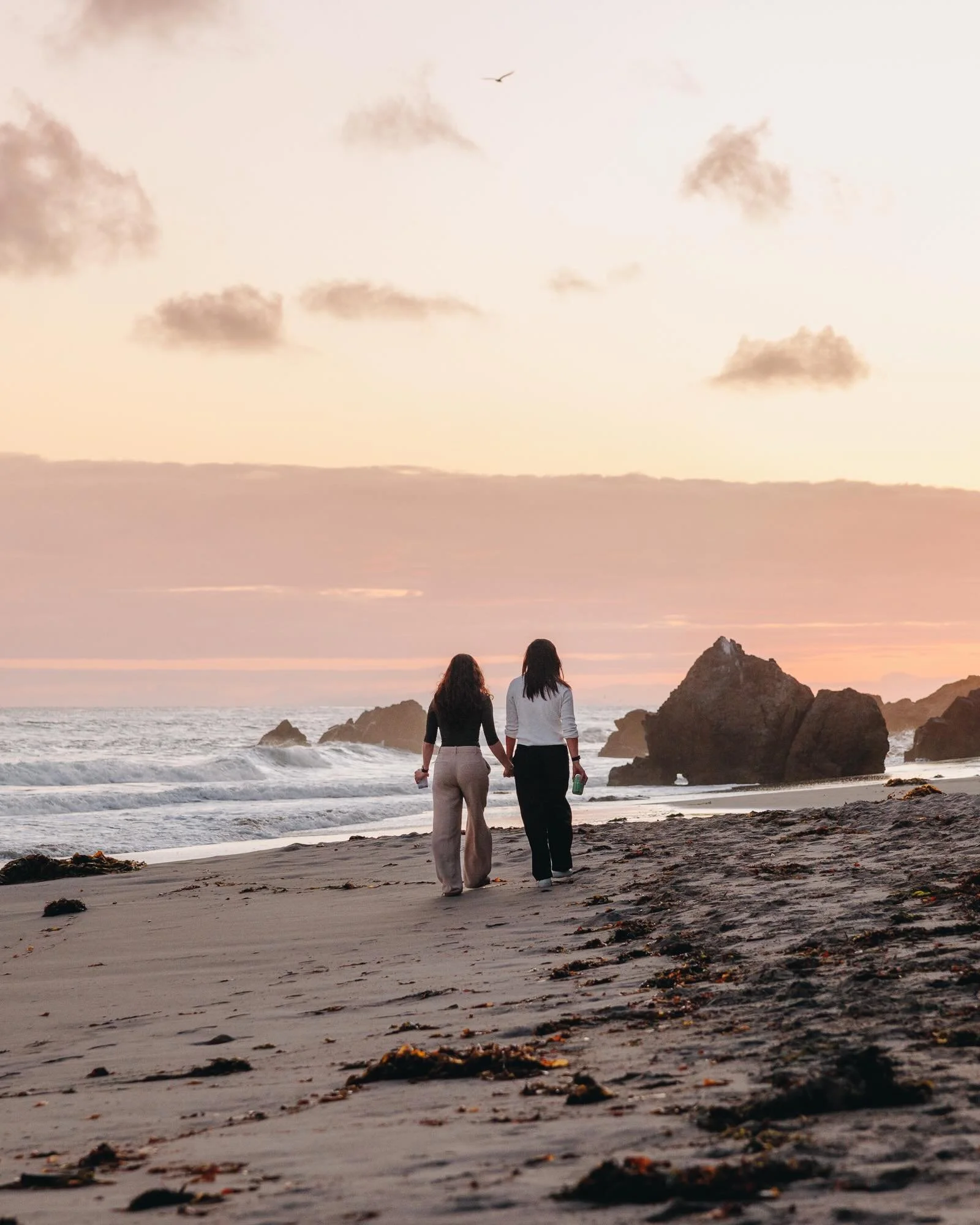 Two women walking on a beach during sunset or sunrise, holding hands, with rocks and ocean waves in the background, cloudy sky, and bird flying in the sky.