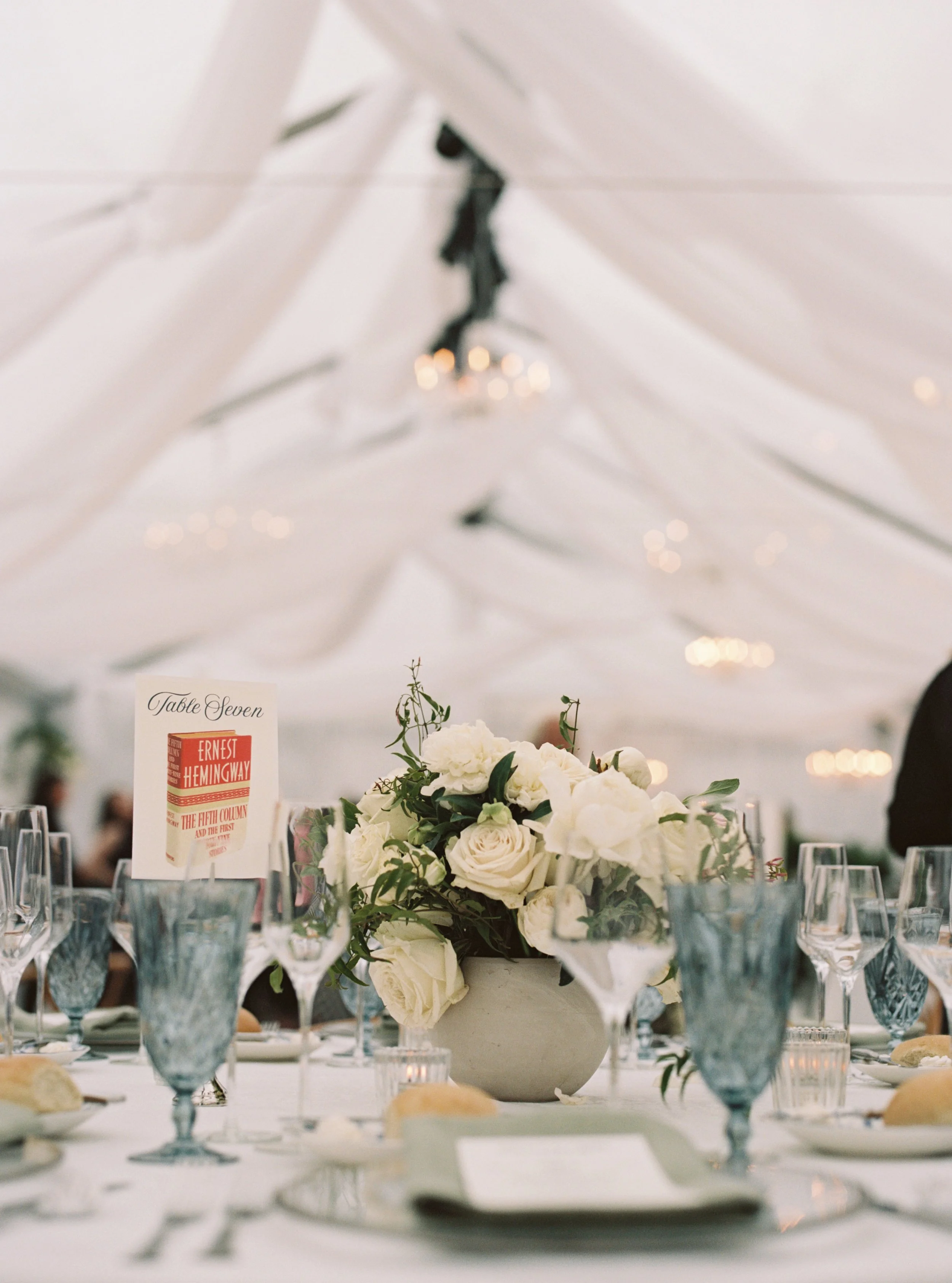 A table decorated with a floral centerpiece of white roses and greenery, surrounded by multiple glasses and a sign indicating it is for guest 'Table Seven', with a blurred chandelier hanging from a white ceiling in a venue with elegant decor.