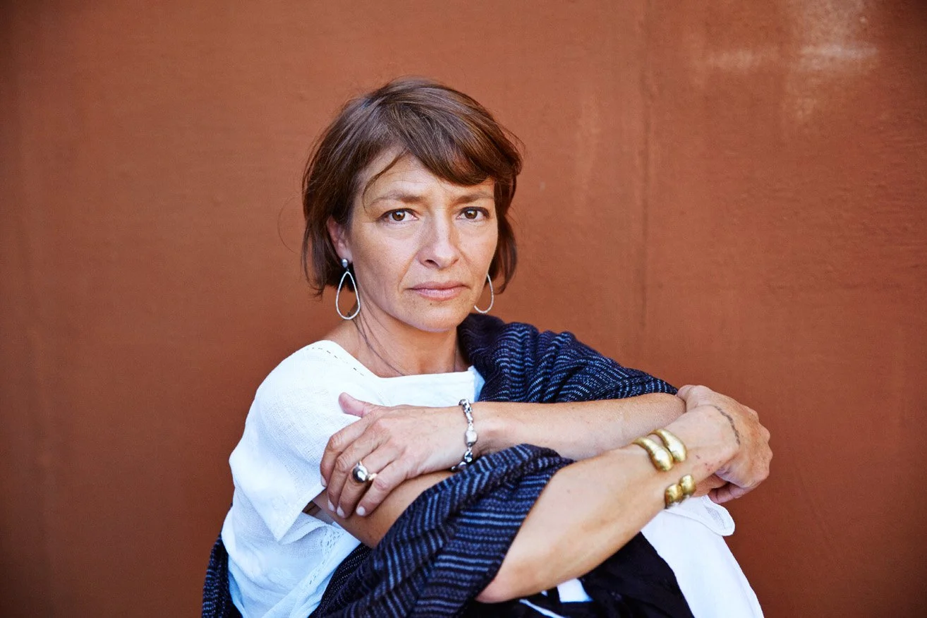 A woman with short brown hair wearing hoop earrings, a white top, and gold jewelry, sits against a brown wall with her arms crossed over her knees, looking directly at the camera.