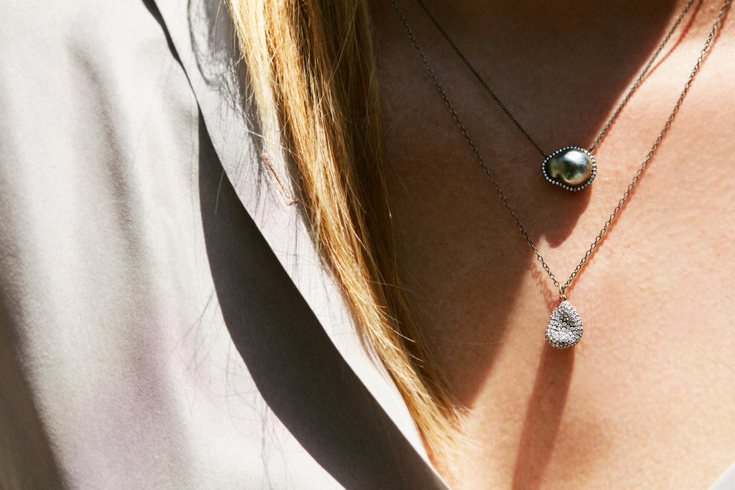 Close-up of a person wearing layered silver necklaces with pendants, and wearing a light-colored blazer over a low-cut top.