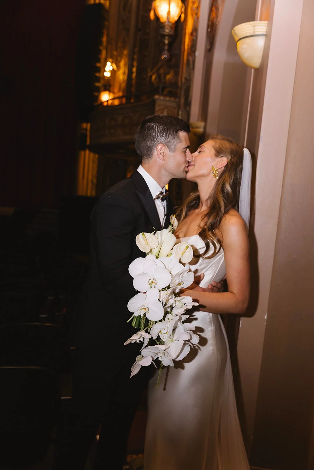 A bride and groom sharing a kiss at their wedding, with the groom in a black tuxedo holding a bouquet of white orchids, and the bride wearing a strapless white gown with long, wavy hair and gold earrings.