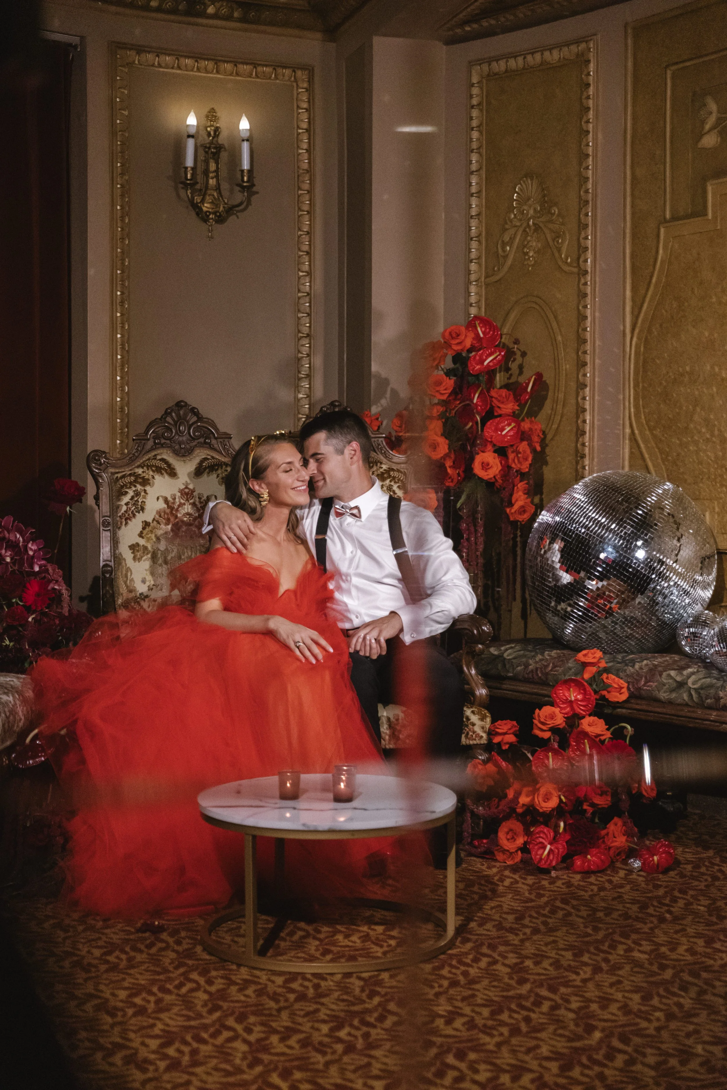 A woman in a red dress and a man in a white shirt and suspenders are sitting close together on a vintage ornate sofa, smiling and embracing in an elegant, gold-decorated room. There are red flowers and a disco ball on the side.