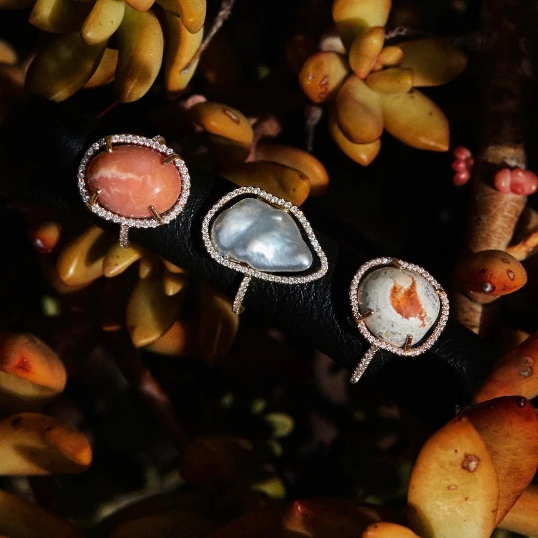 Three rings with larger stones, one peach-colored, one iridescent, and one white with orange markings, each surrounded by small diamonds, set on a black band with a background of orange and brown succulent plants.
