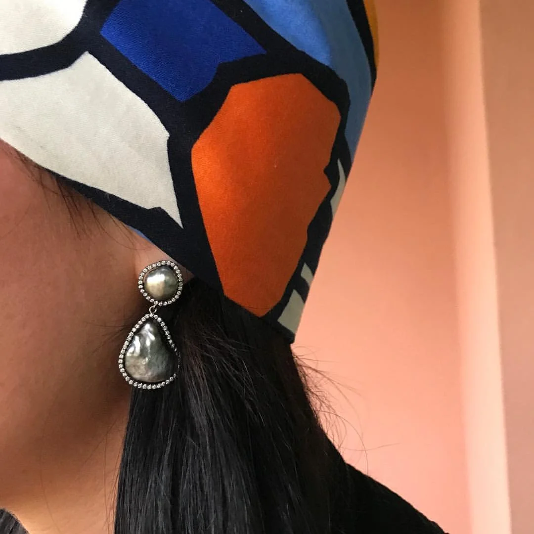 Close-up of a person's ear wearing large pearl earrings with rhinestones, and a colorful geometric-patterned headscarf in orange, blue, white, and black.