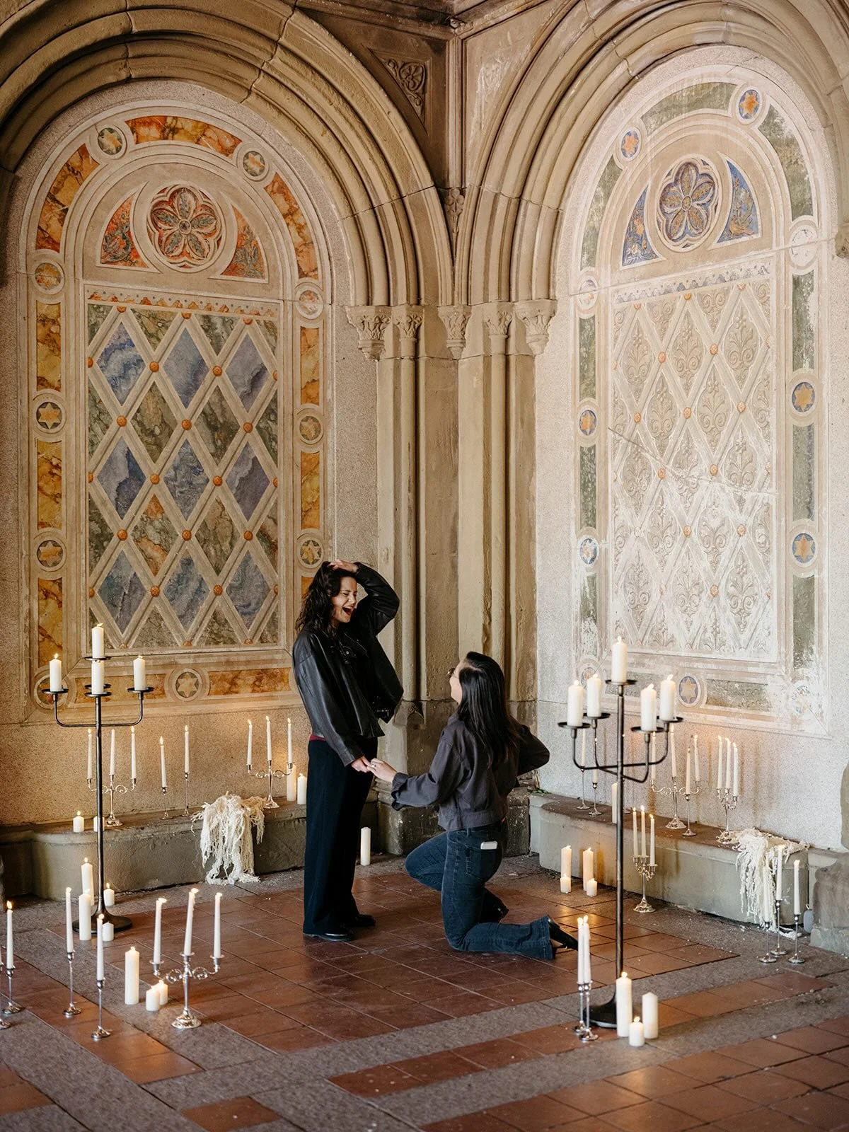 A woman proposing marriage to a woman inside a church with stained glass windows, surrounded by lit candles.