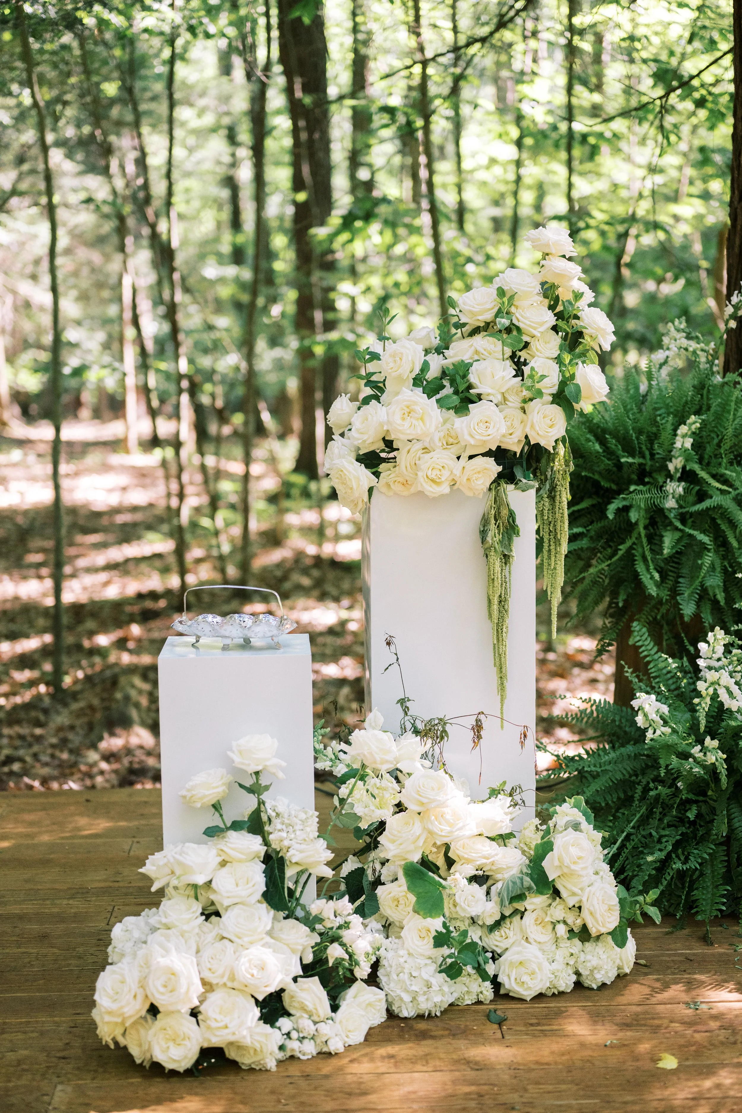 White roses arranged on a white stand and cascading onto a wooden floor, surrounded by green foliage in a forest setting.