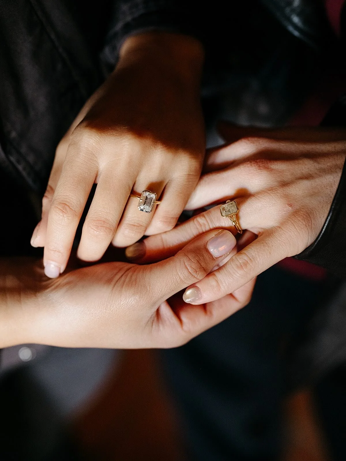 Two hands holding each other, each wearing a ring. The left hand has a large rectangular clear gemstone ring, while the right hand has a smaller gold ring with a square gemstone. The background is dark.