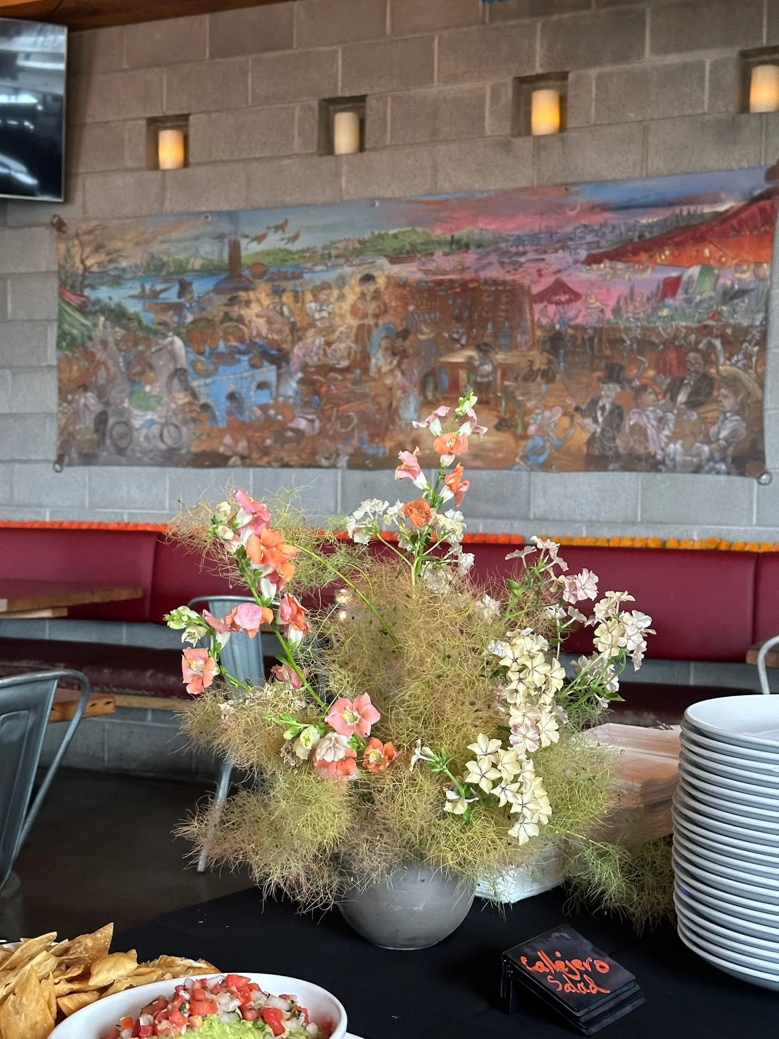 A large floral arrangement with pink, white, and orange flowers and greenery is on a table in front of a gray wall with a mural depicting a lively marketplace scene and a parking lot with cars.