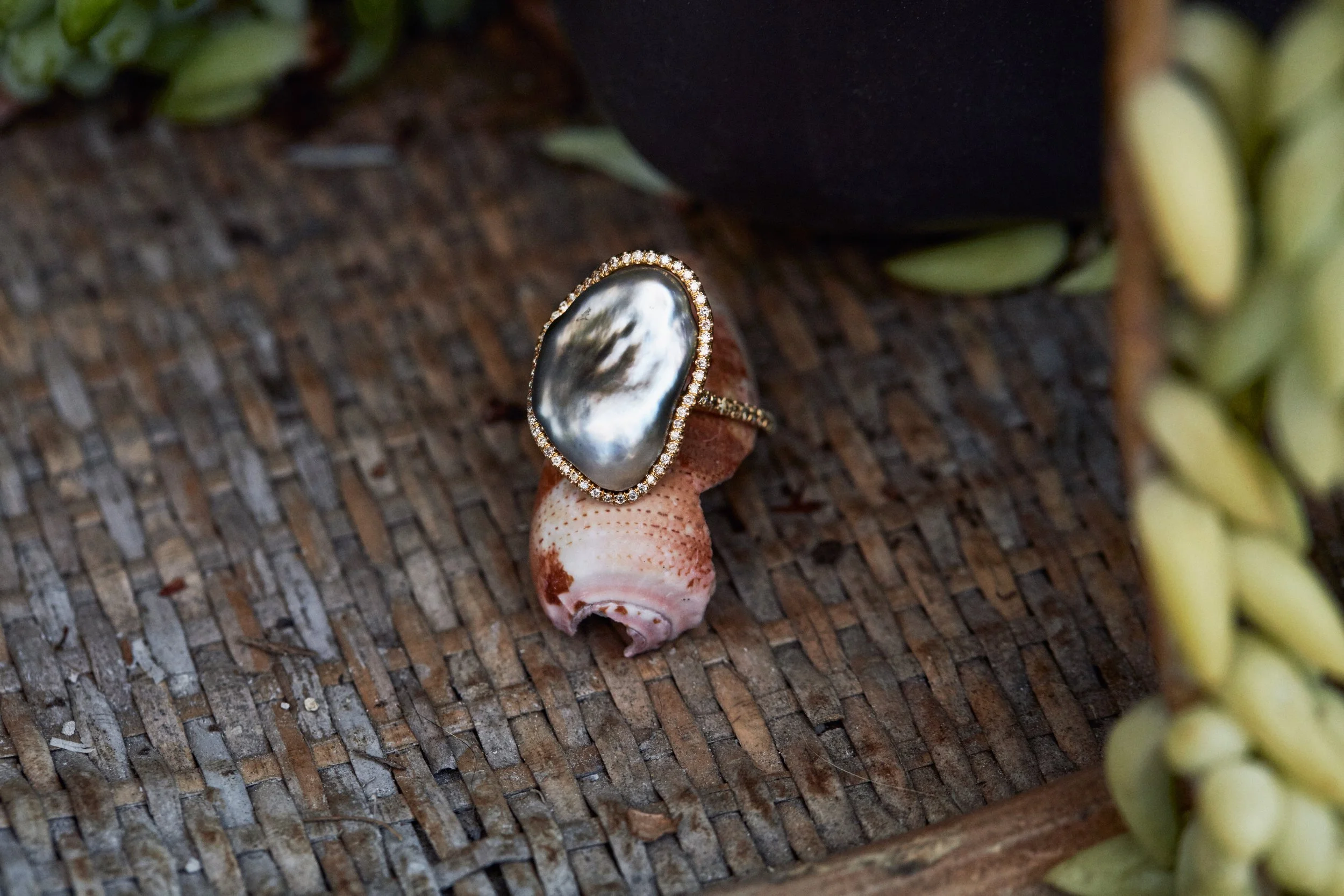 A large pearl ring with a diamond border on the band, resting on a person's finger with shell and succulent plants in the background.