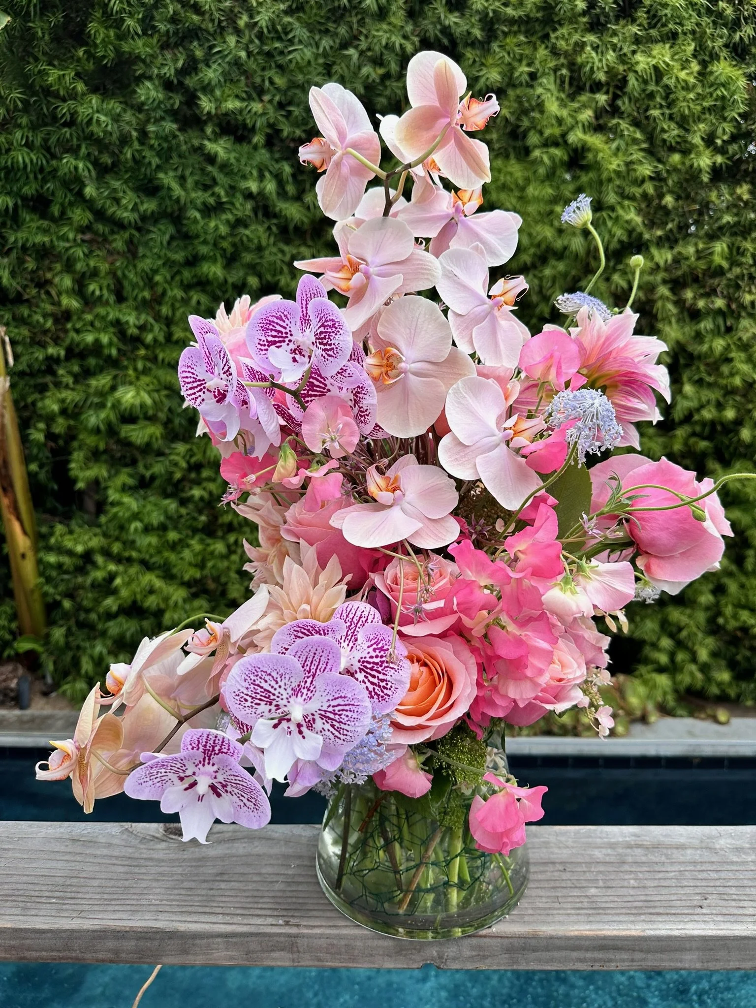 A glass vase filled with pink, purple, and white flowers, including orchids and roses, placed on a wooden surface with a green leafy background.