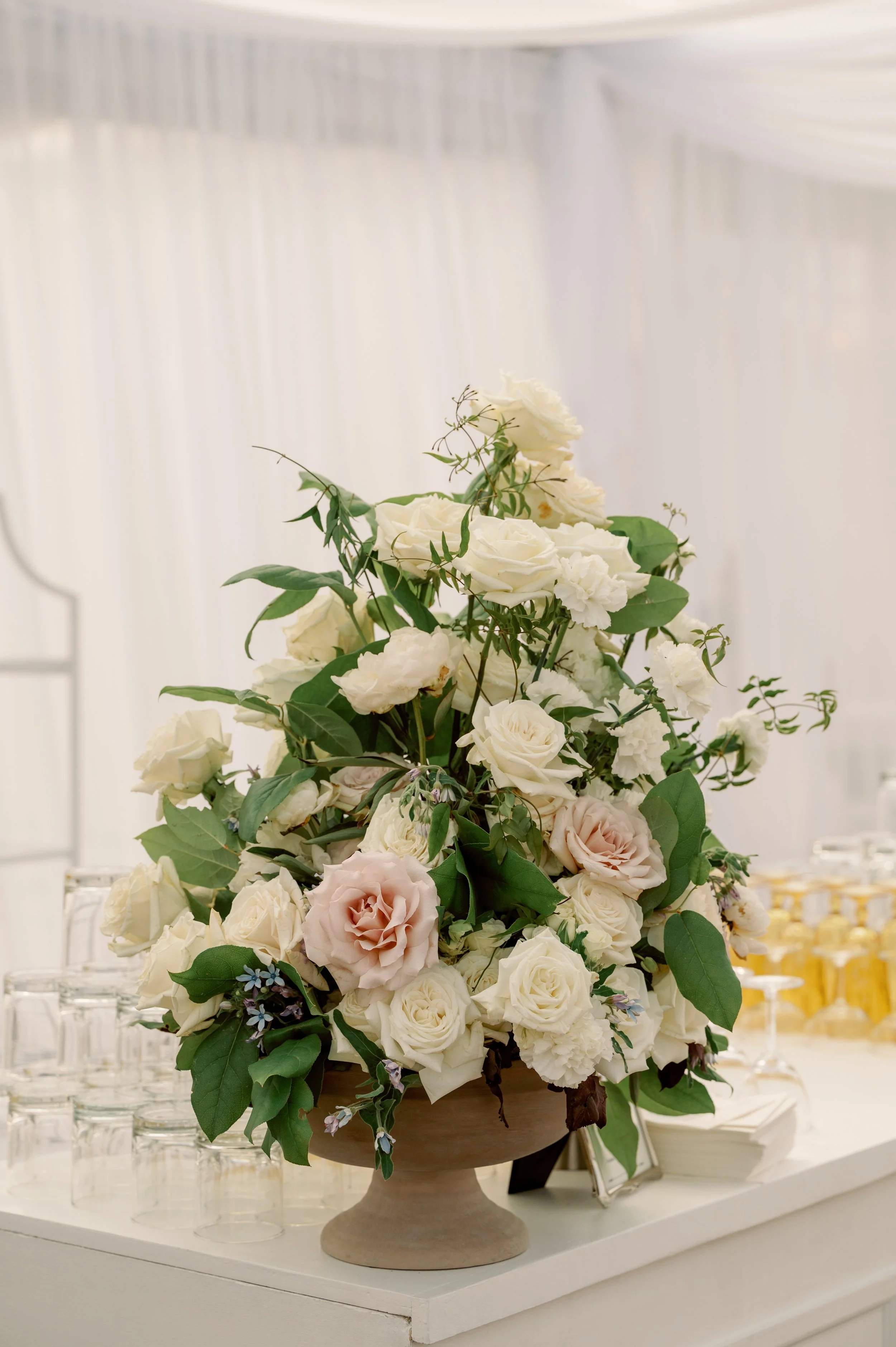 A large bouquet of white and pale pink roses and greenery in a beige pedestal vase at an event.
