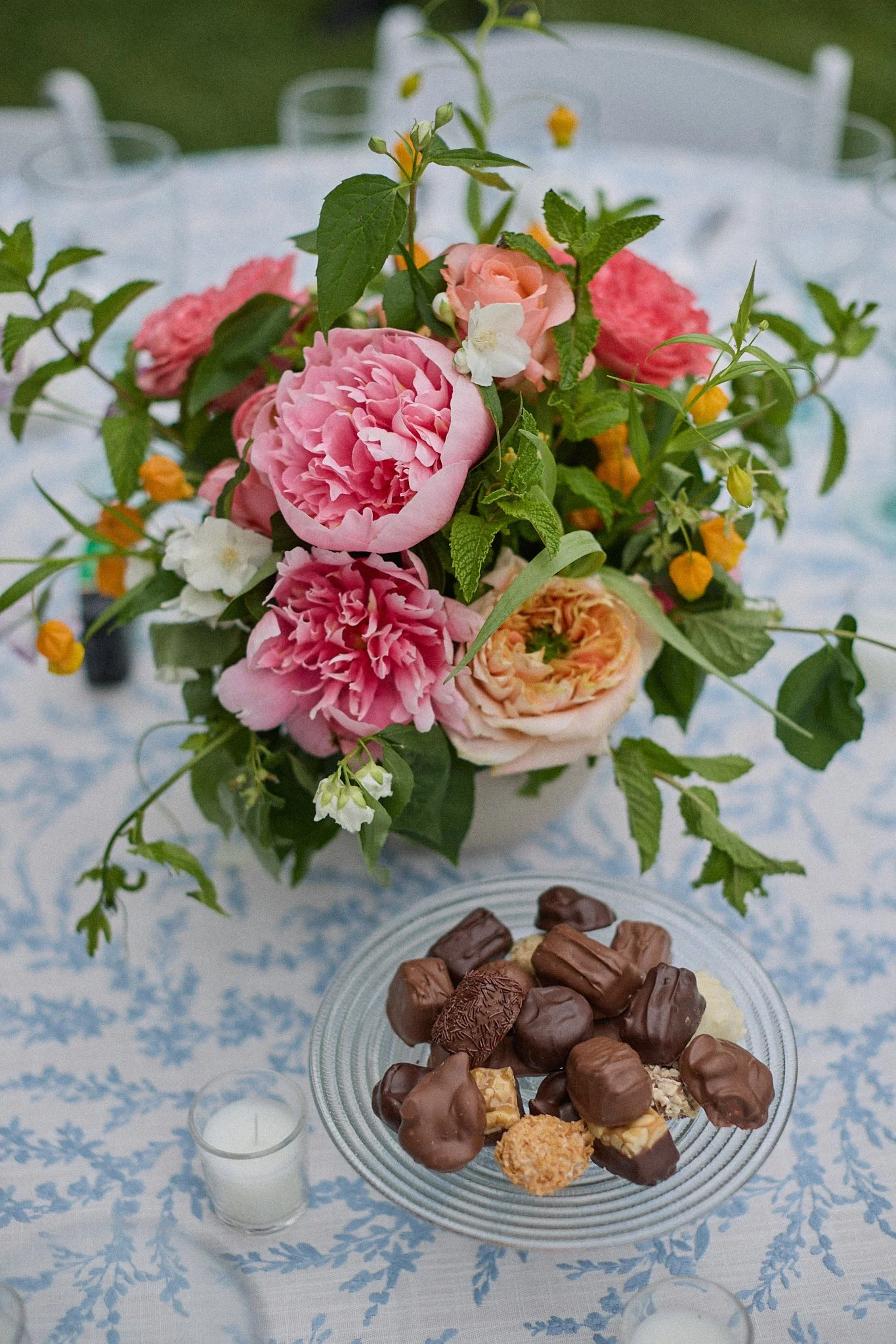 A bouquet of pink, orange, white, and yellow flowers with green leaves on a table draped with a blue and white patterned cloth, with a glass bowl of assorted chocolates and two small white candles nearby.