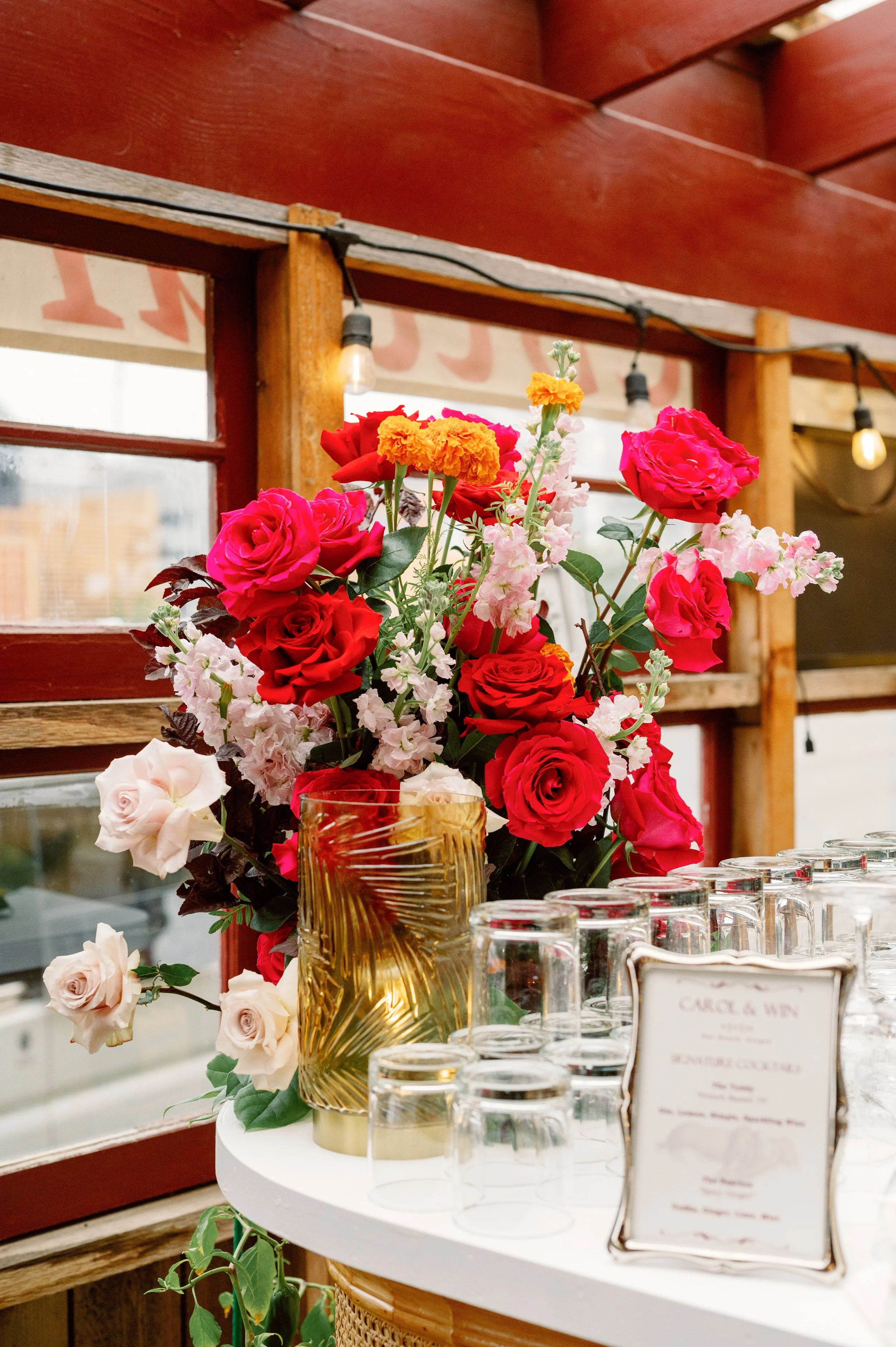A floral arrangement with pink, red, orange, and white flowers on a white round table, with glasses and a menu, inside a rustic venue with wooden beams and string lights.
