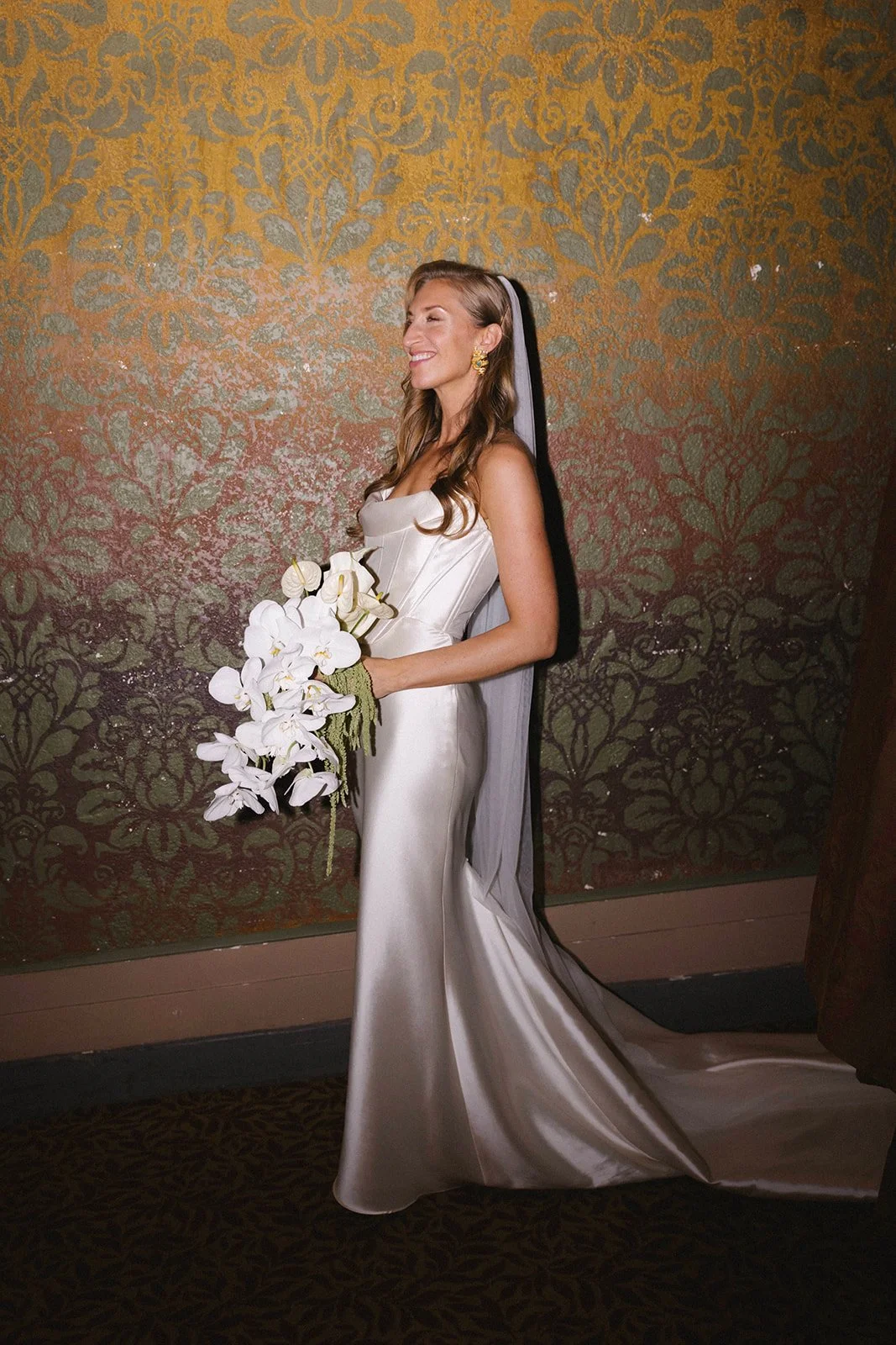 A woman in a satin wedding dress with a veil holding a bouquet of white orchids, standing against a decorative gold and brown wall, smiling with eyes closed.