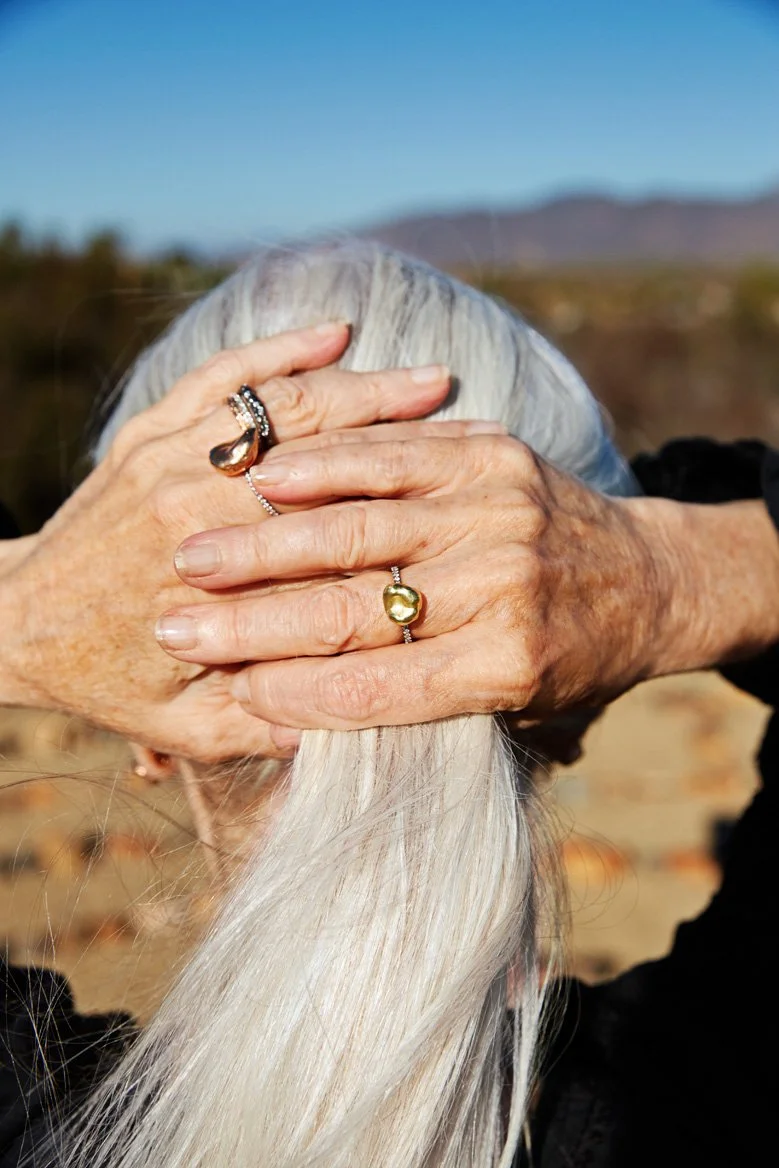 Older woman with silver hair holding her head with both hands, outdoors with a clear blue sky and desert landscape in the background.