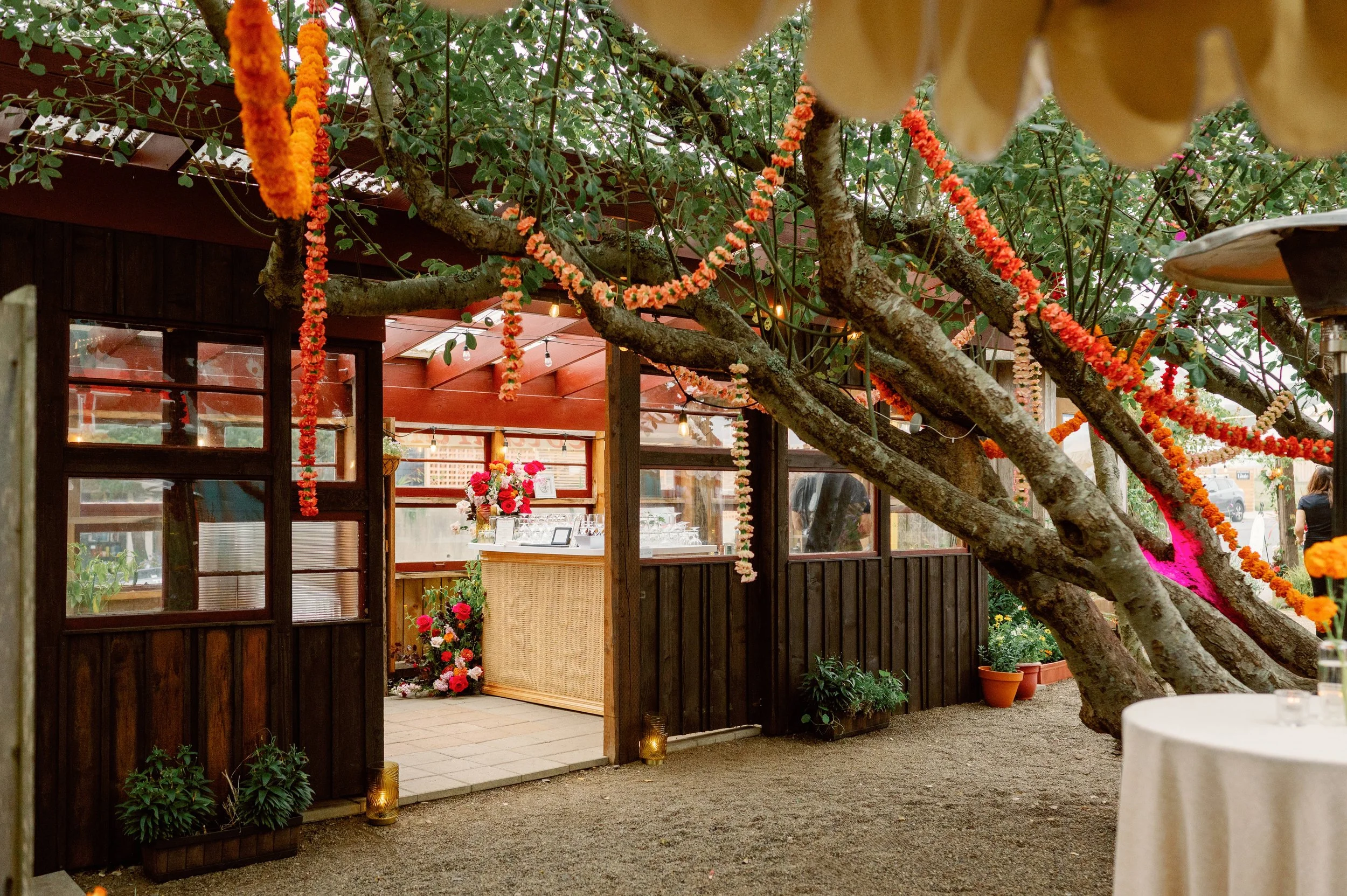 Decorated outdoor venue with string lights, orange and pink floral garlands, potted plants, and a table with flowers inside a wooden structure, set up for a celebration or event.