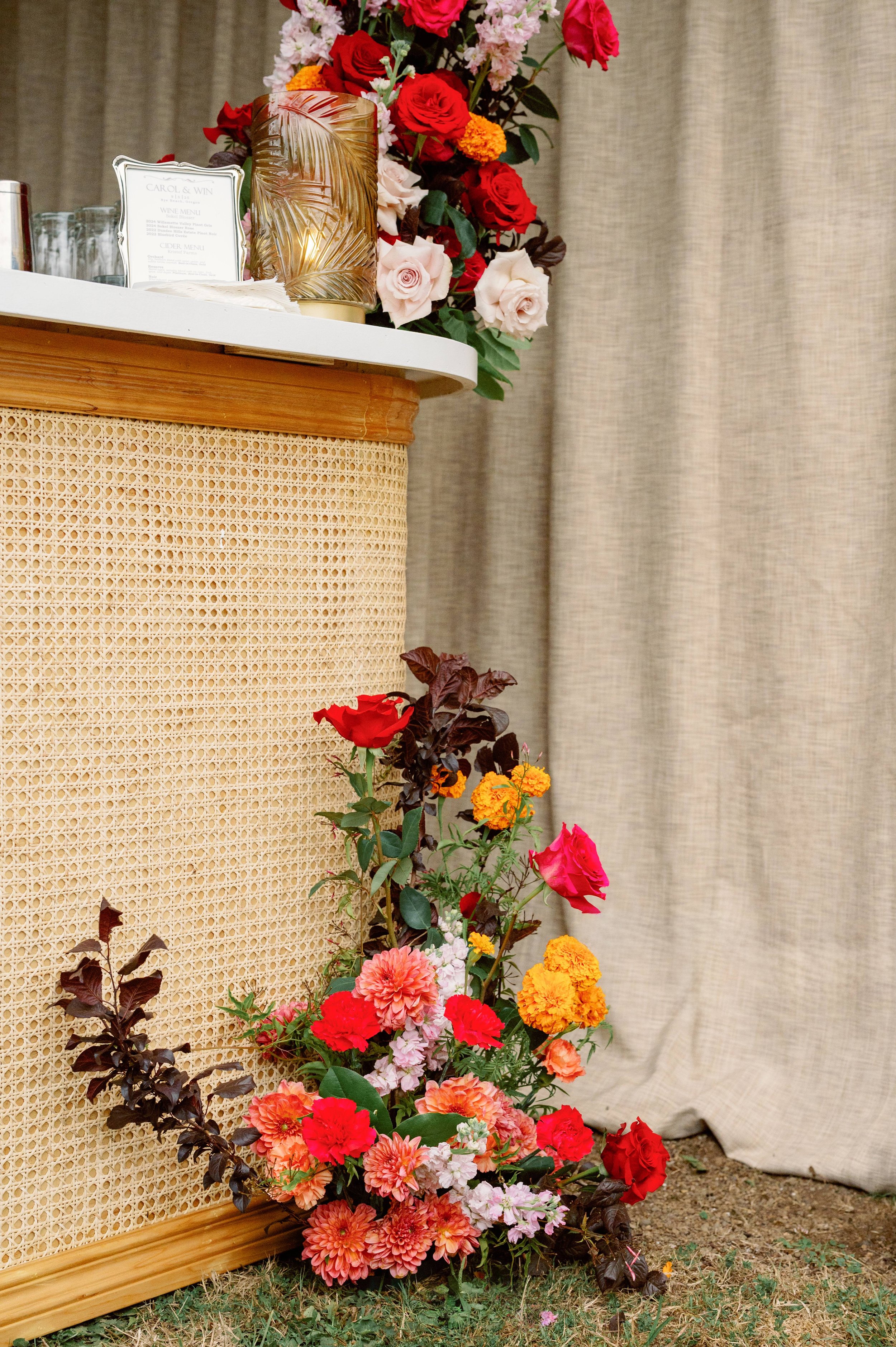 A floral arrangement with roses, marigolds, dahlias, and other flowers cascading beside a beige rattan panel, with a white marble countertop and a tall, gold decorative vase nearby, set against a neutral beige curtain background.