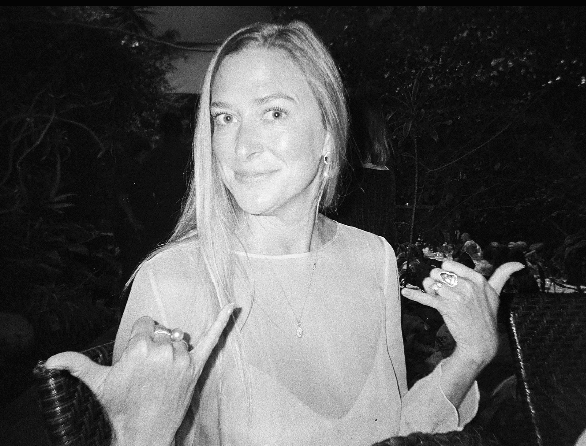 Black and white photo of a woman with long blonde hair smiling and making rock and roll hand gestures with both hands, wearing rings and earrings, outdoors at night with trees and other people in the background.