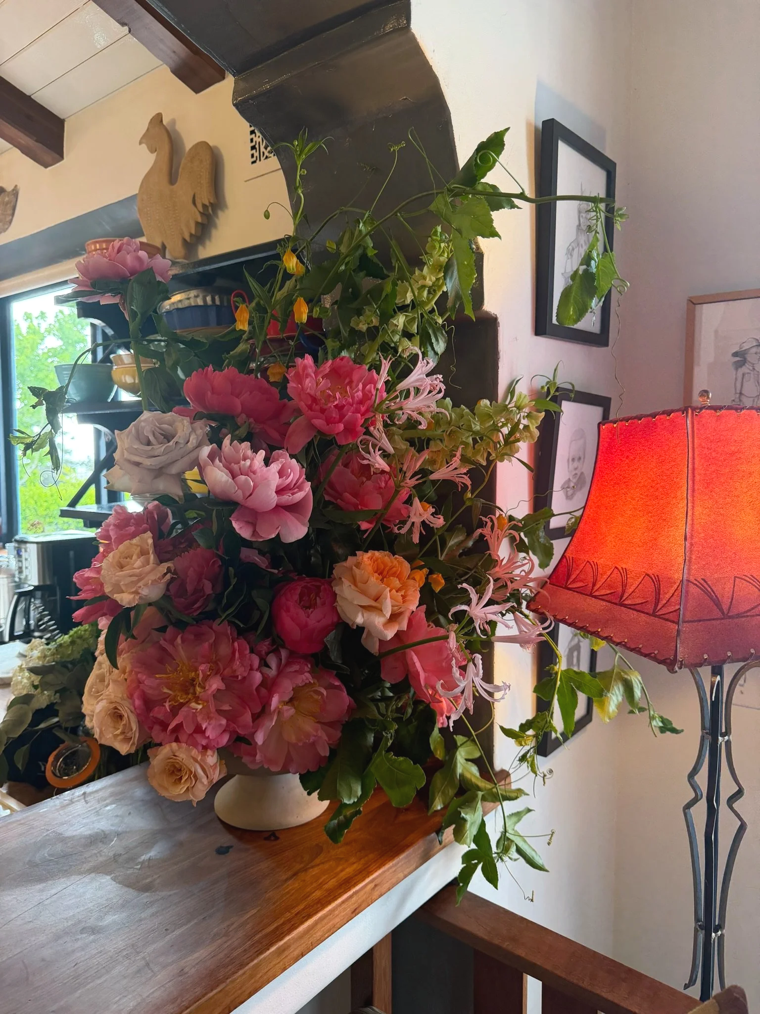 A bouquet of pink, white, and yellow flowers on a wooden table near a window, with a red lampshade in the background and framed artwork on the wall.