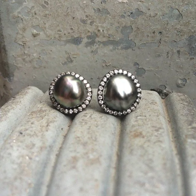 Pair of black pearl earrings with a surrounding circle of small diamonds, placed on a textured gray surface.