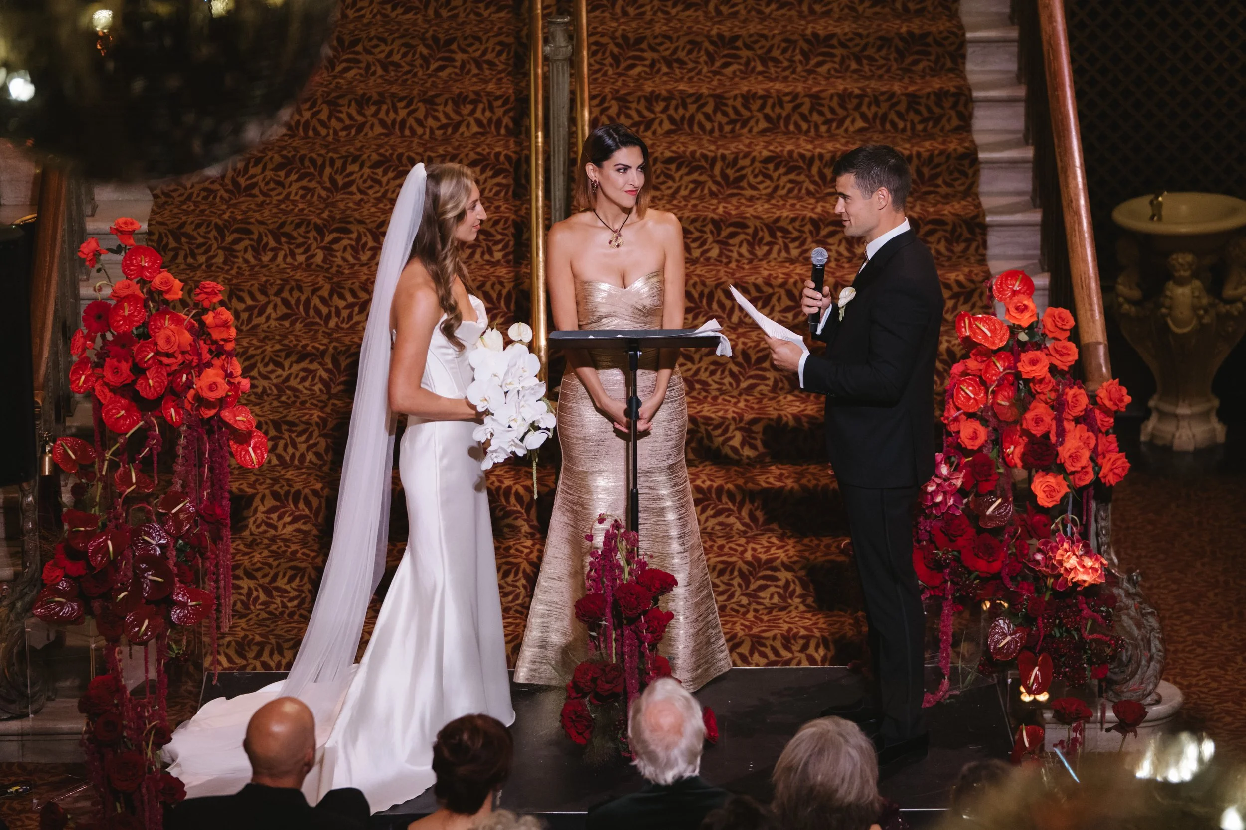 A wedding ceremony with a bride and groom standing before a officiant, surrounded by red and pink floral decorations, on a staircase with a patterned red carpet, with guests seated and observing.
