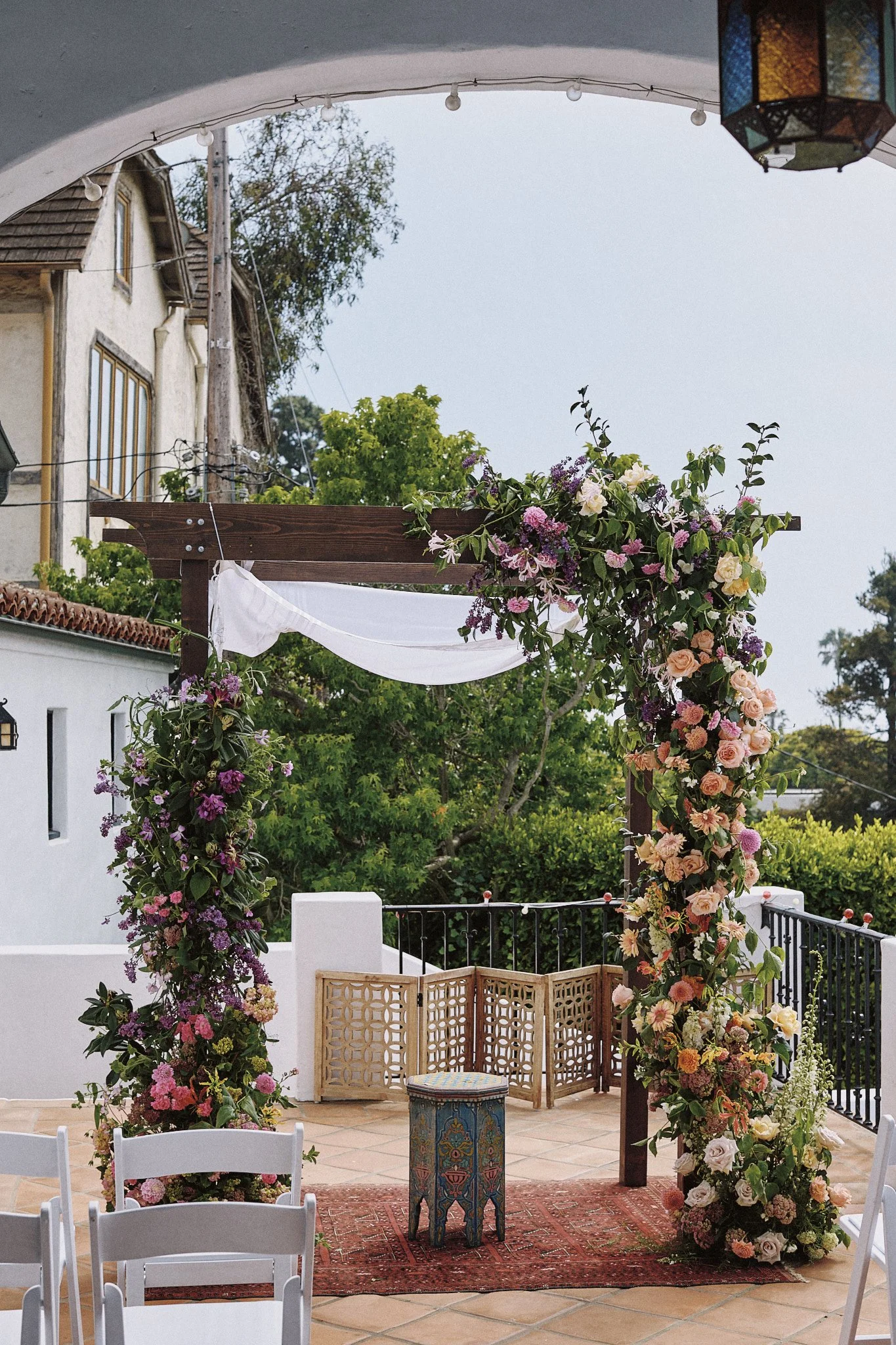Outdoor wedding ceremony setup with an arch decorated with a cascade of pink, purple, and white flowers, a red Persian rug, white folding chairs, and a small intricately patterned table, against a backdrop of greenery and a white building.