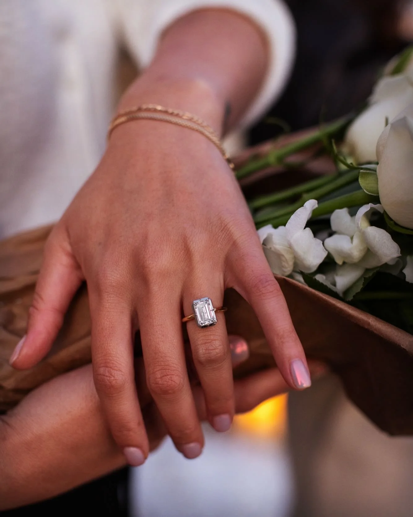 Close-up of a woman's hand showing an emerald-cut ring with a large rectangular gemstone, possibly a diamond, on her ring finger. The hand has a braided bracelet on the wrist and is placed over a basket with white flowers.