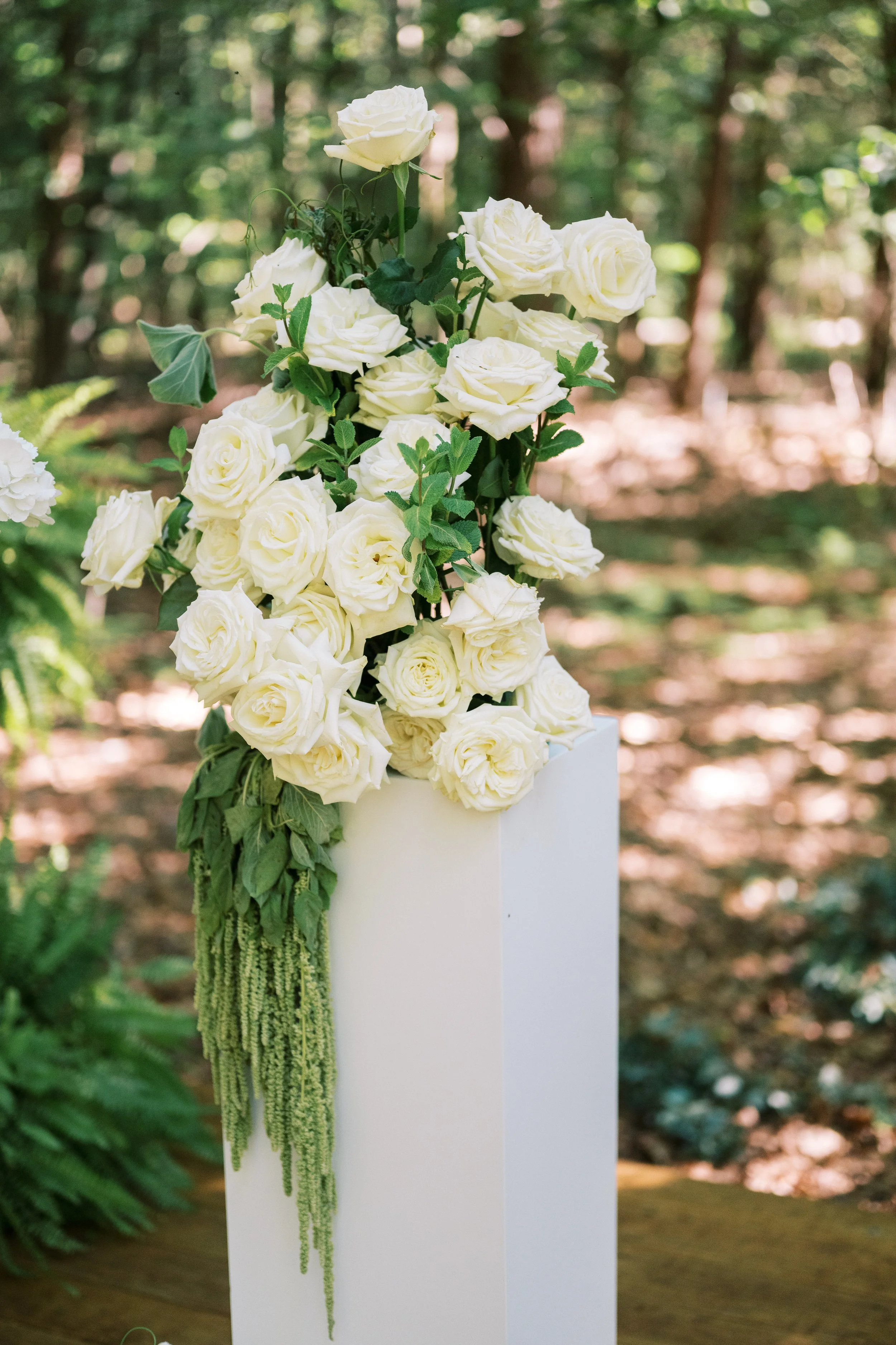 A bouquet of white roses and green foliage with hanging greenery, arranged on a white pedestal outdoors with a blurred wooded background.