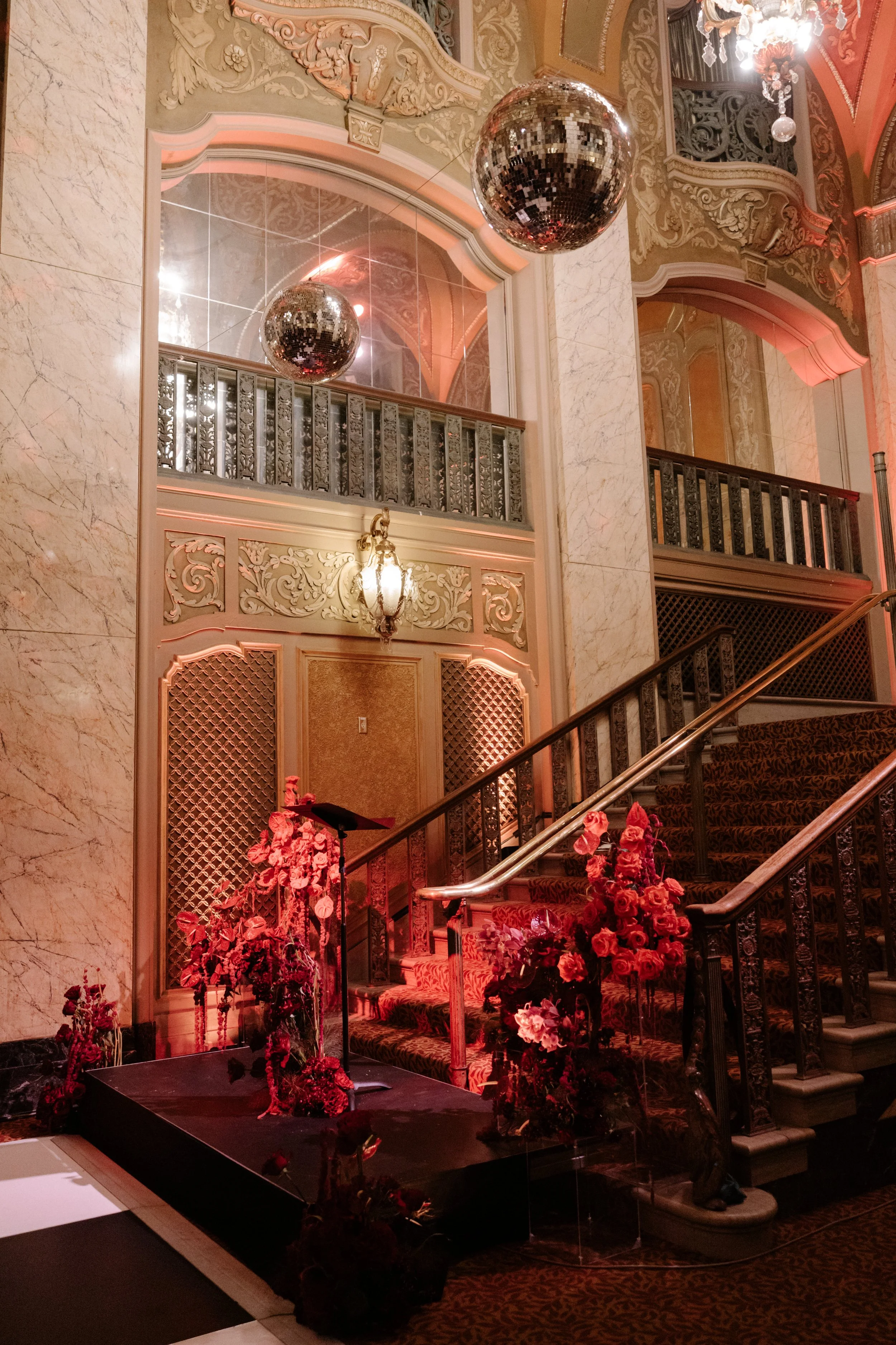 Elegant interior with ornate decorations, a marble staircase adorned with red and pink flowers, a mirror reflecting a disco ball, and vintage-style lighting.
