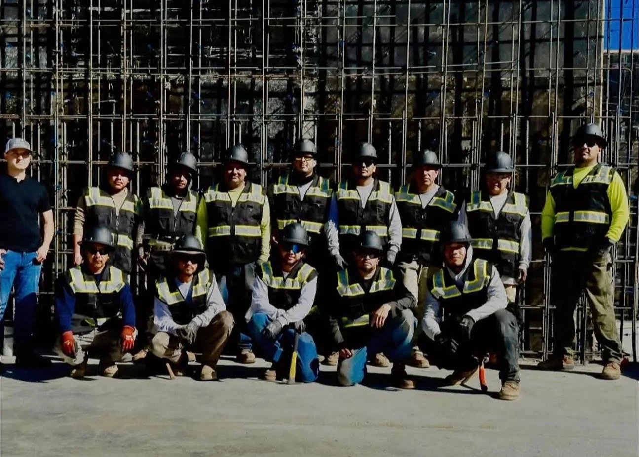 Group of construction workers wearing safety gear, including helmets and vests, posing in front of a steel rebar structure at a construction site.