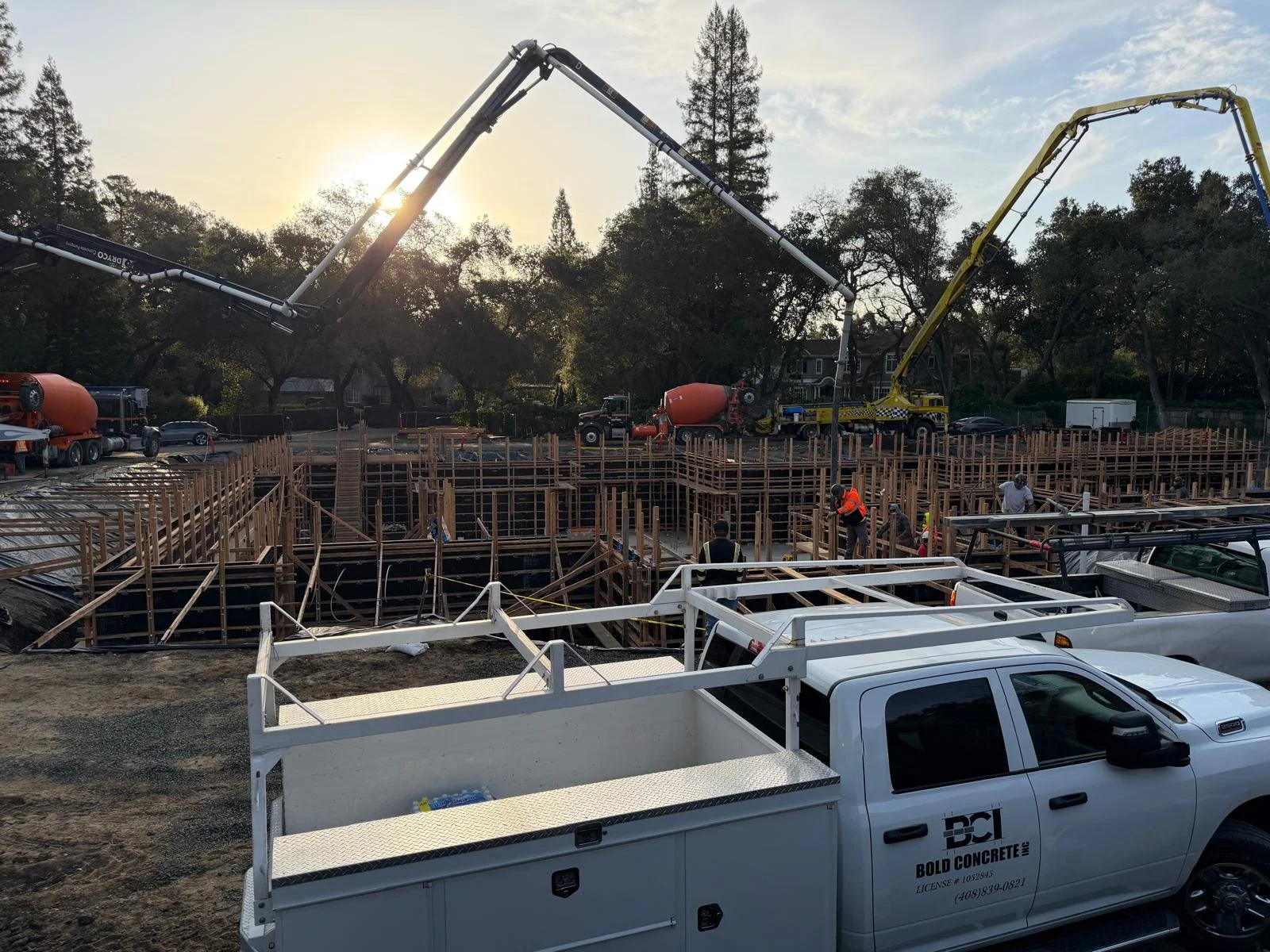 Construction site with wooden framework, construction workers, concrete mixers, and trucks, under a sunset sky with trees in the background.