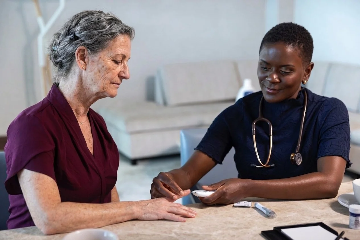 Private healthcare nurse and professional for aging parents in the Aventura /Hollywood/ Fort Lauderdale area wearing a stethoscope, showing a device to an elderly woman at a table in a medical setting.