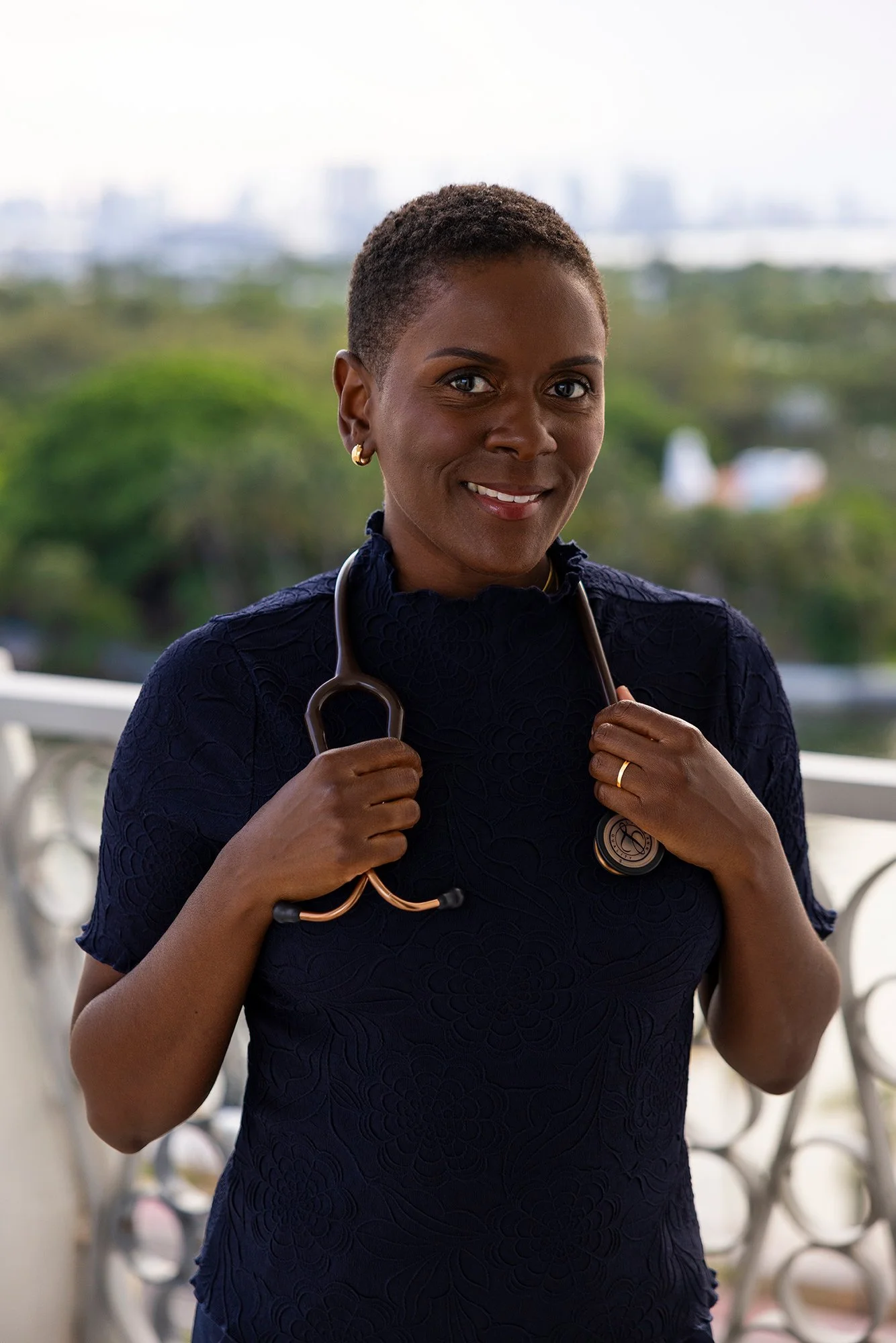 A smiling female healthcare professional holding a stethoscope, standing outdoors with a blurred cityscape and greenery in the background. Private nurse for aging parents Aventura /Hollywood/ Fort Lauderdale
