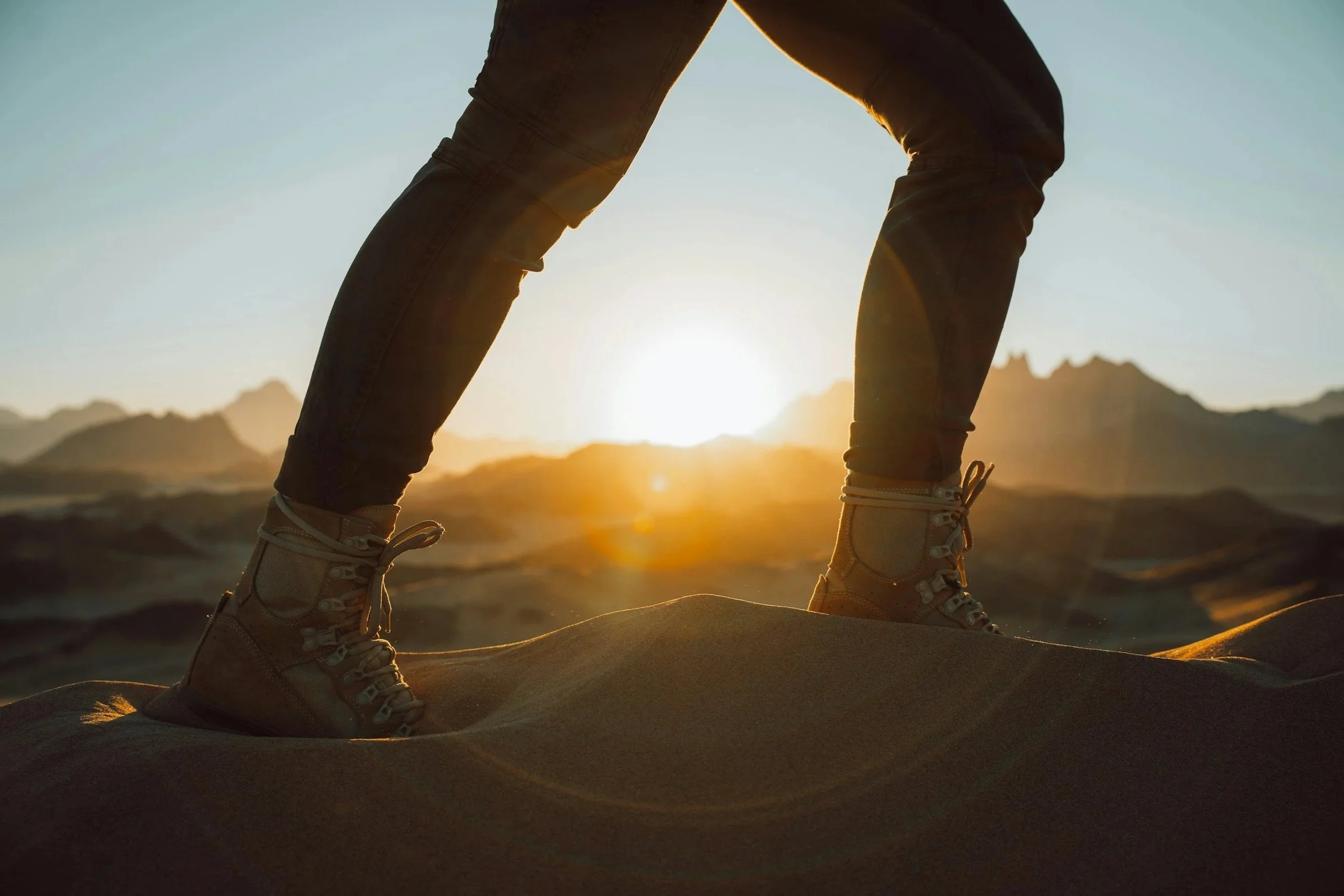 Close-up of a person's legs in hiking boots standing on sand dunes at sunset.