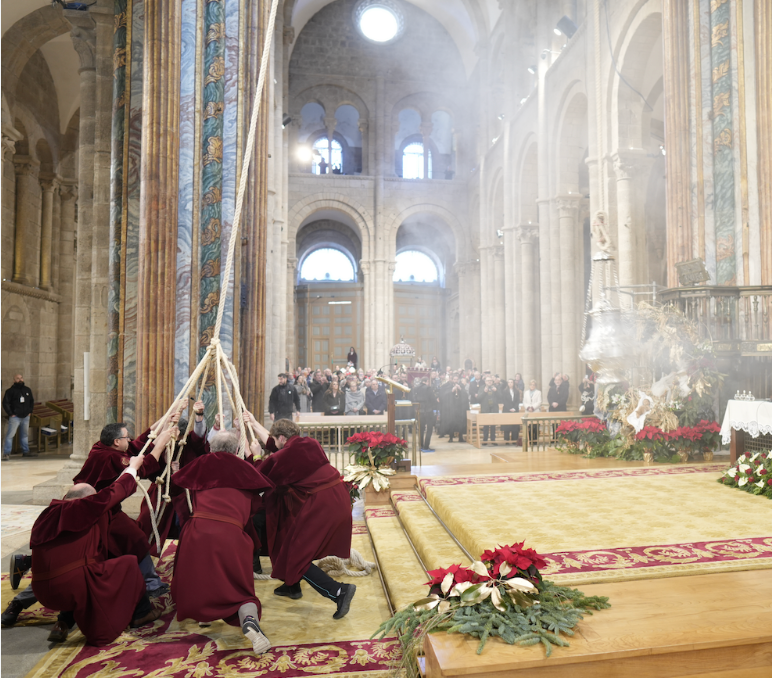 Eight tiraboleiros in red robes pulling ropes to swing the Botafumeiro inside Santiago Cathedral, with pilgrims gathered near the altar.
