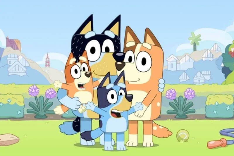 Four animated dogs from Bluey TV show standing outdoors in a garden, hugging each other, with houses, trees, and flowers in the background.