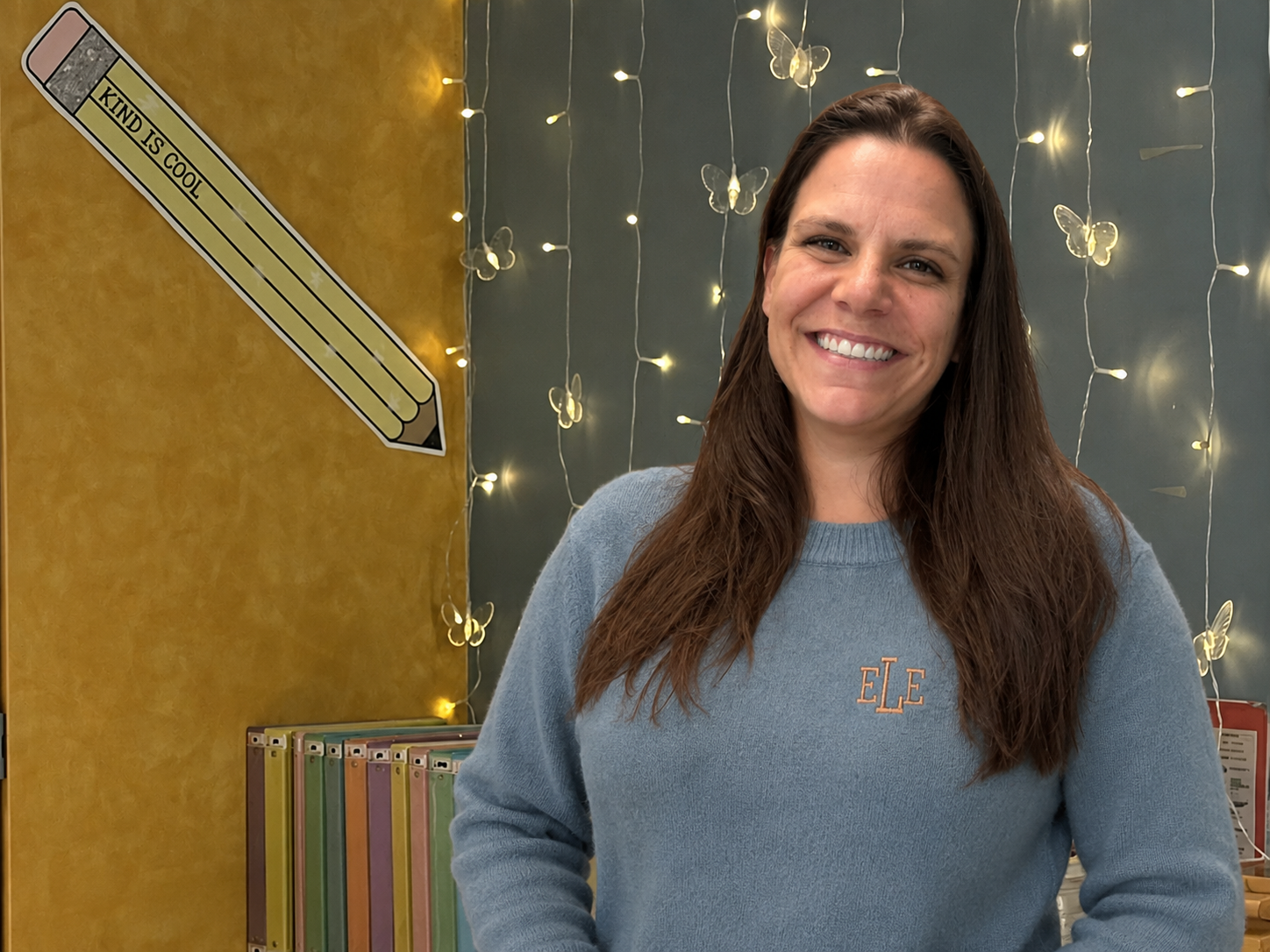 A woman with long brown hair smiling in front of a decorated wall with fairy lights and butterflies; a large pencil cutout with 'Kind is Cool' written on it is on the wall, and colorful folders are on a shelf.