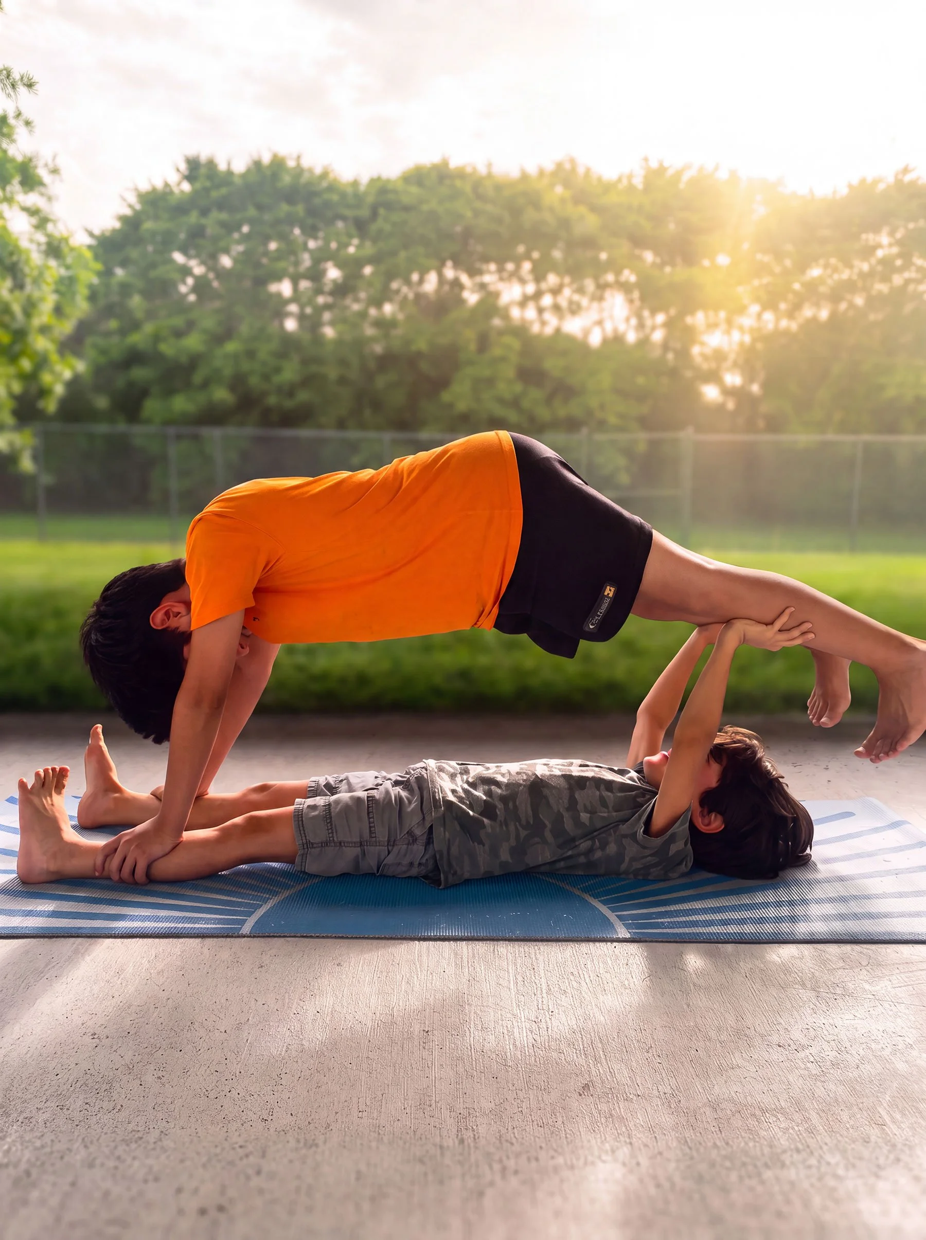Children practicing yoga outdoors on a mat with trees and sunlight in the background, forming a human pyramid.