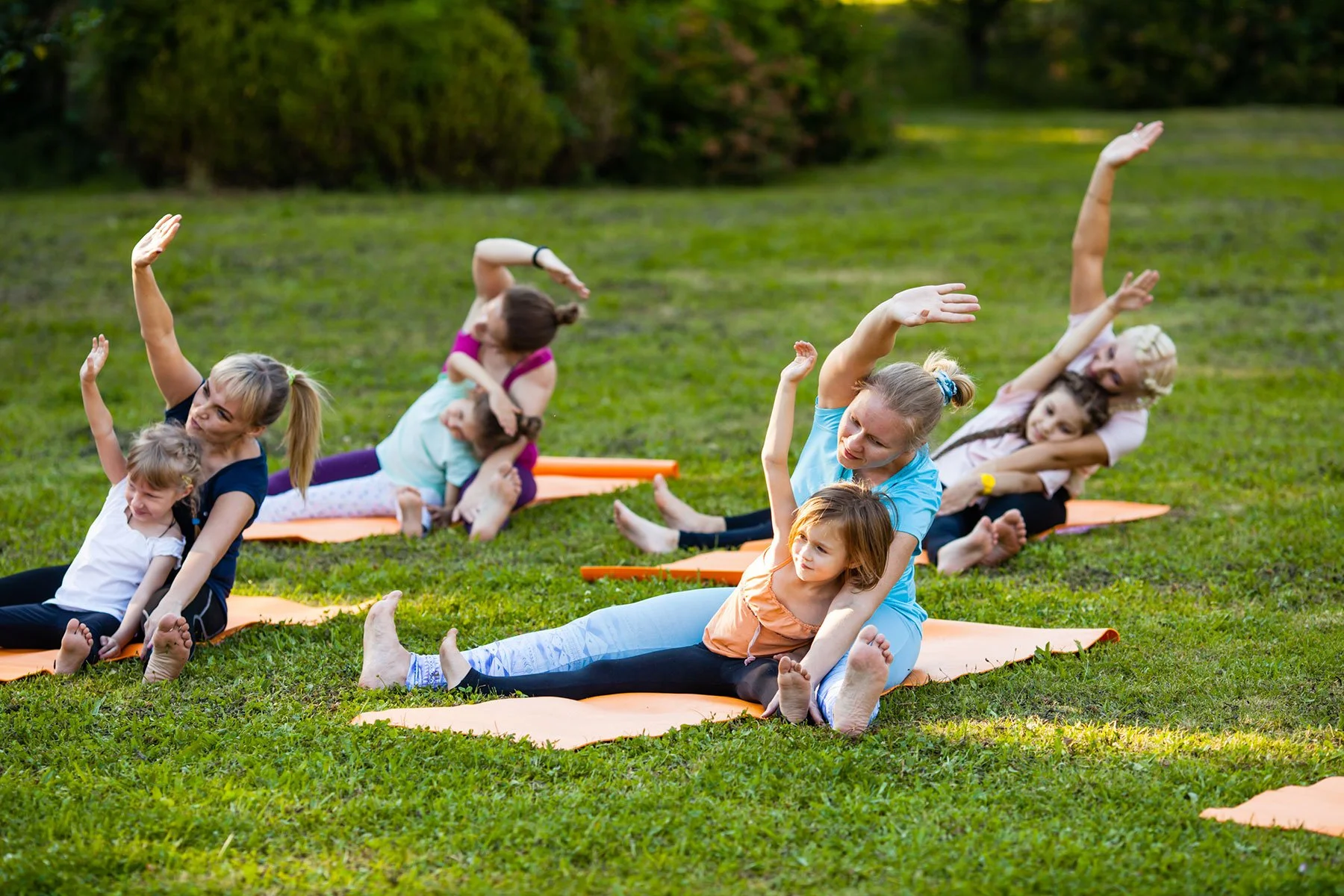 Group of children and adults practicing yoga outdoors on yoga mats in a grassy park, with trees in the background.