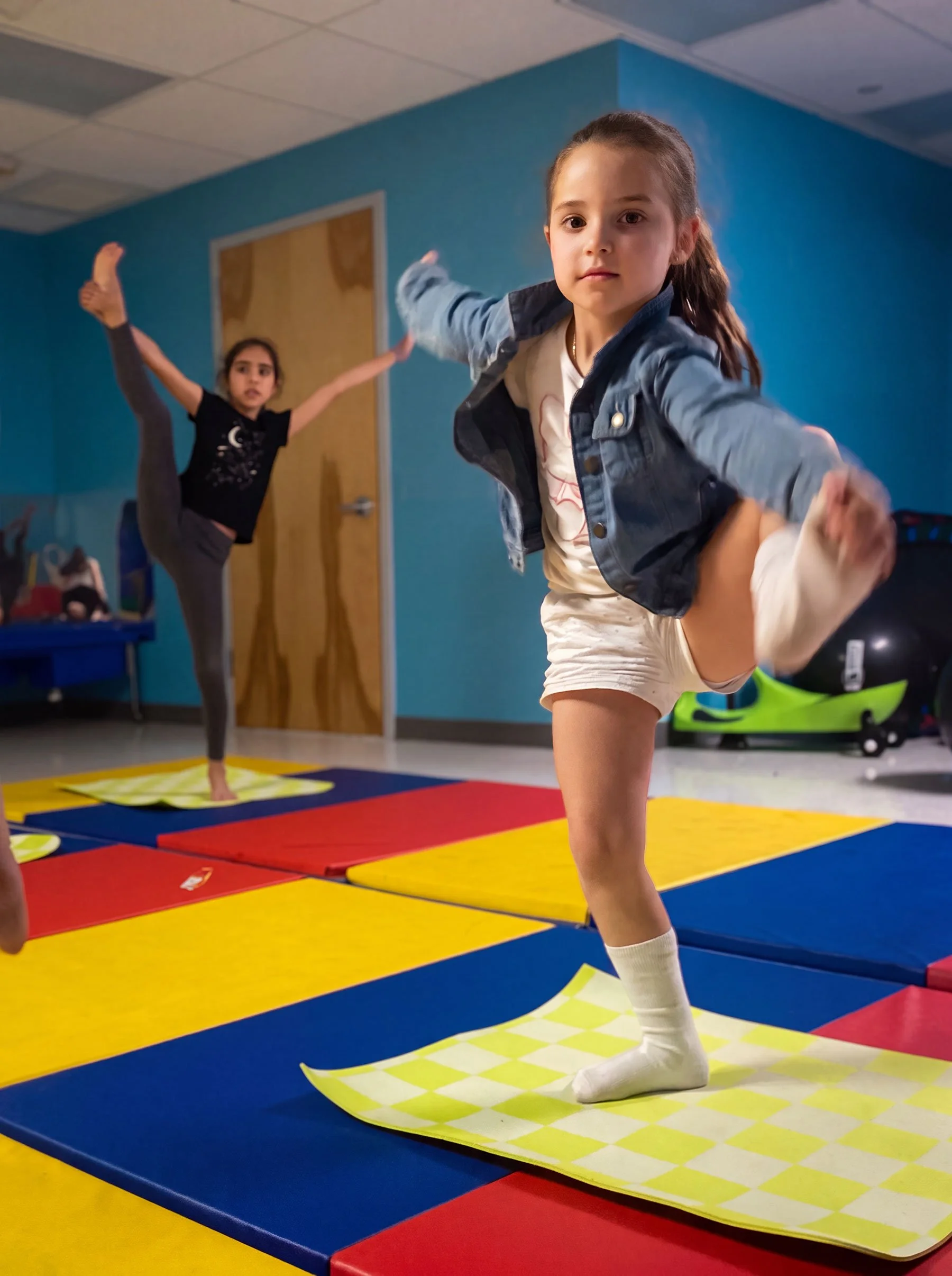 Two young girls practicing gymnastics in a colorful indoor gym, one in the foreground balancing on a yellow mat with a checkered pattern, the other in the background doing a high kick on a blue and red padded mat.