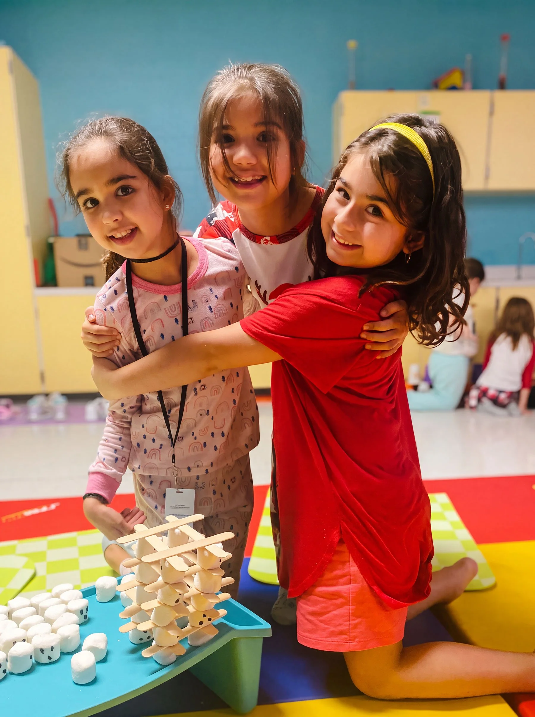 Three young girls smiling and hugging each other while playing with marshmallow and wooden stick game in a playroom with colorful mats and yellow cabinets.