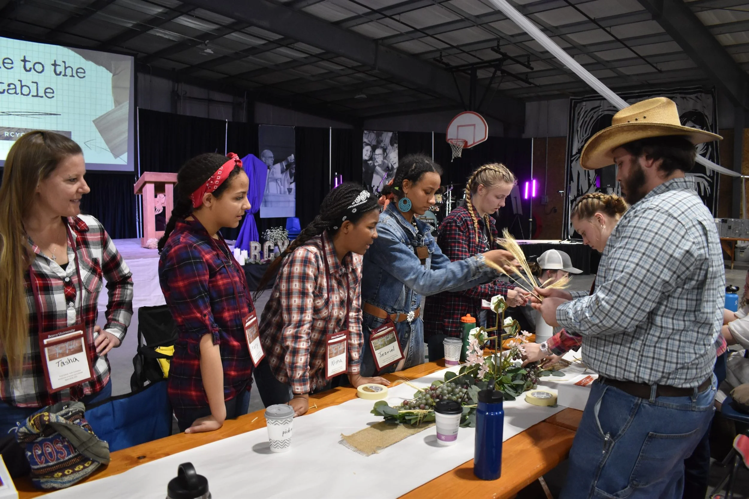 A group of young women and a man participating in a crafting activity at a table during an indoor event. The women are wearing plaid shirts and name tags, and the man is wearing a plaid shirt and a cowboy hat. The table is decorated with flowers and 