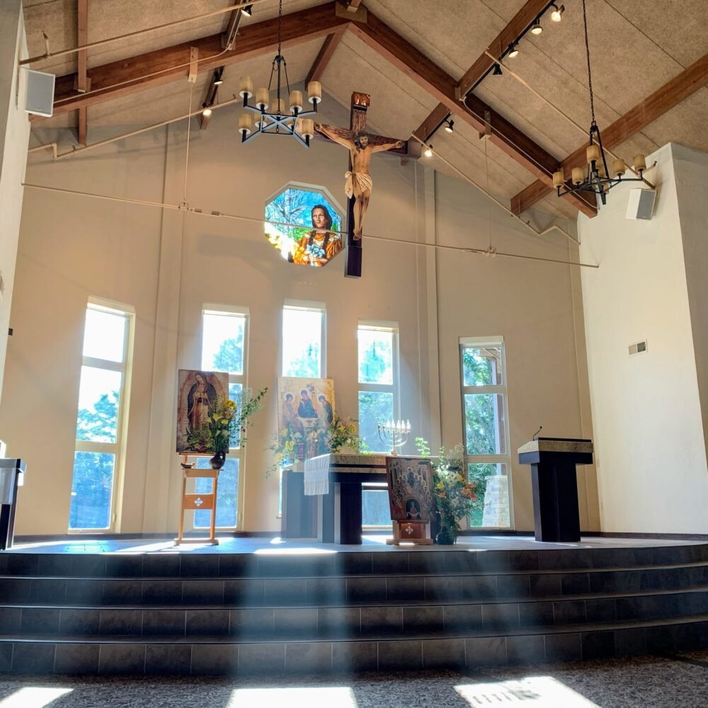 Interior of a church with an altar, religious icons, and a large crucifix hanging from the ceiling. Sunlight streams through tall windows, illuminating the space.