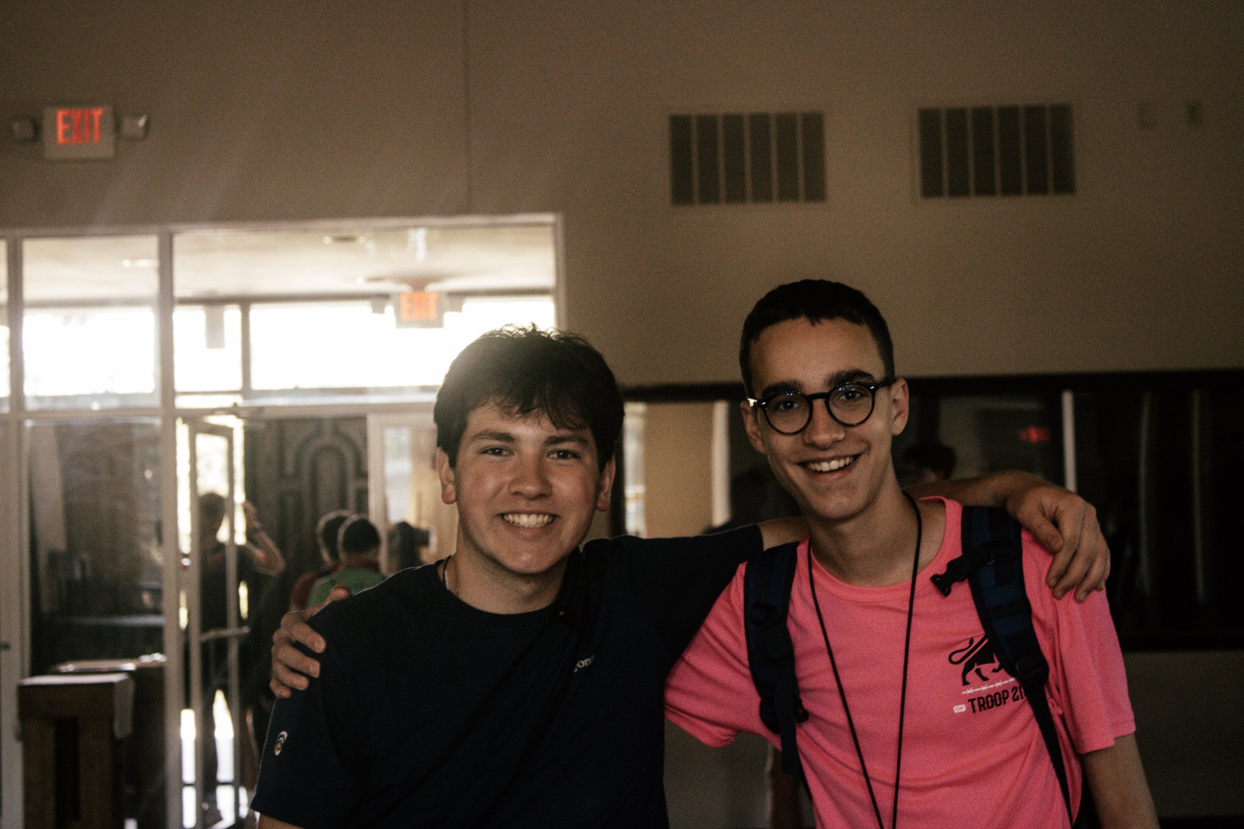 Two young men smiling, with one having his arm around the other, in an indoor setting with mirror and exit signs visible.