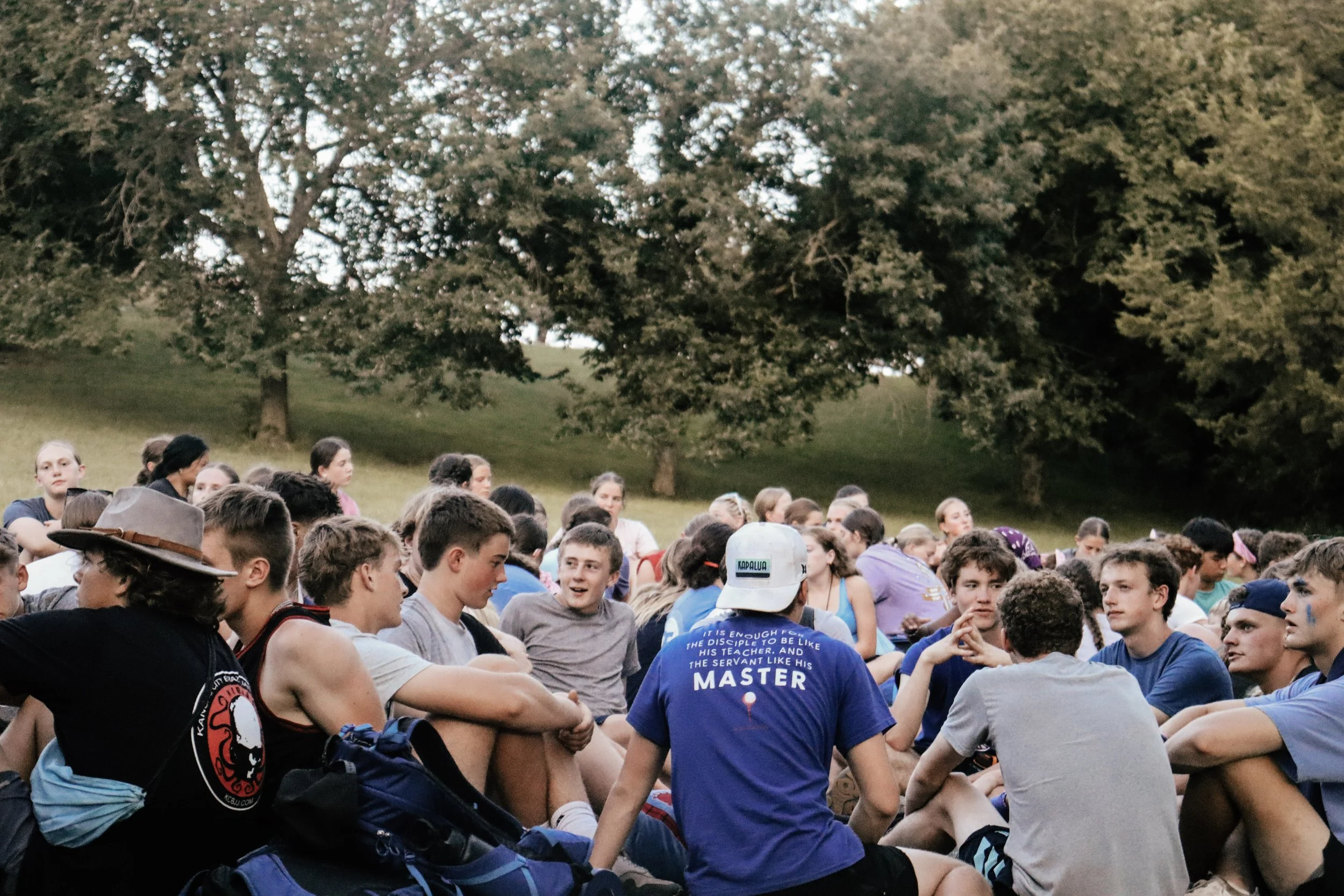 A large group of young people sitting on grass outdoors, surrounded by trees, engaging in conversations and activities on a sunny day.